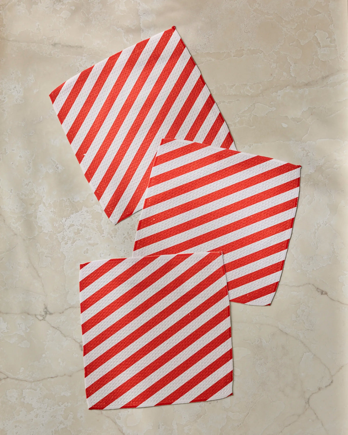 Three napkins from the Geometry Peppermint Lane recycled kitchen towels dishcloth set, featuring red and white stripes, are laid out on a light beige marble surface with each one slightly overlapping the others at different angles.