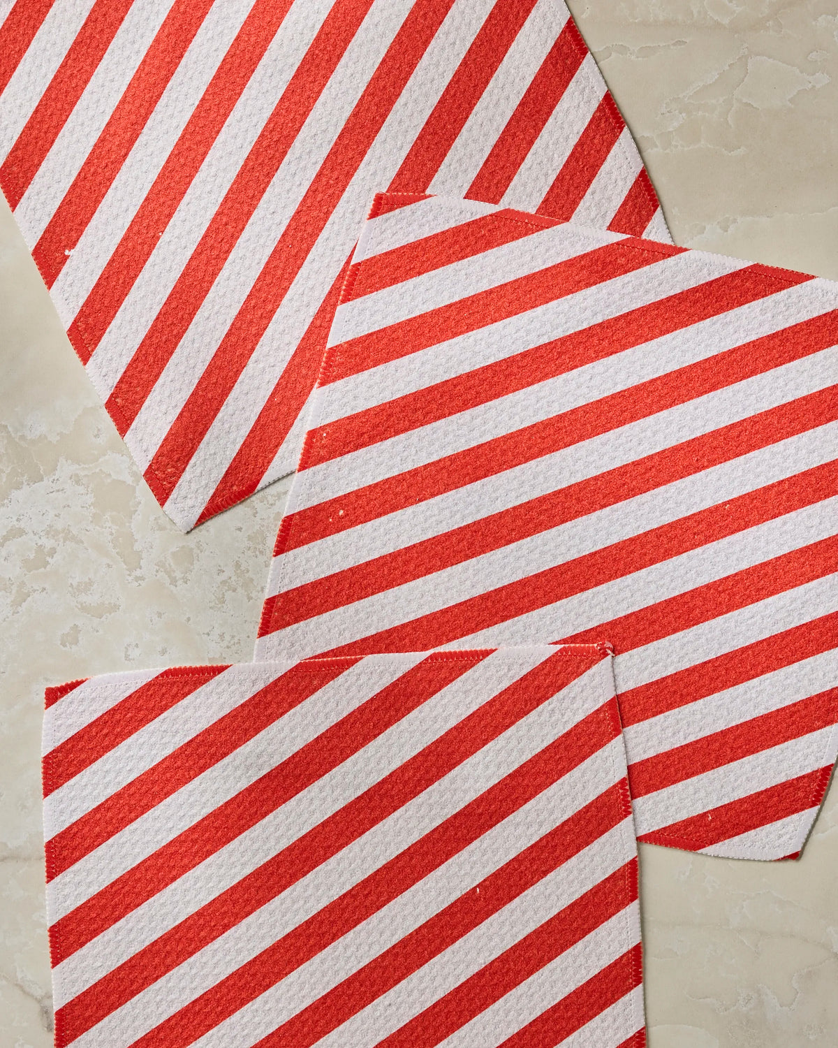 Three white Peppermint Lane napkins by Geometry, featuring diagonal red stripes and crafted from recycled materials, are arranged on a beige, textured surface.