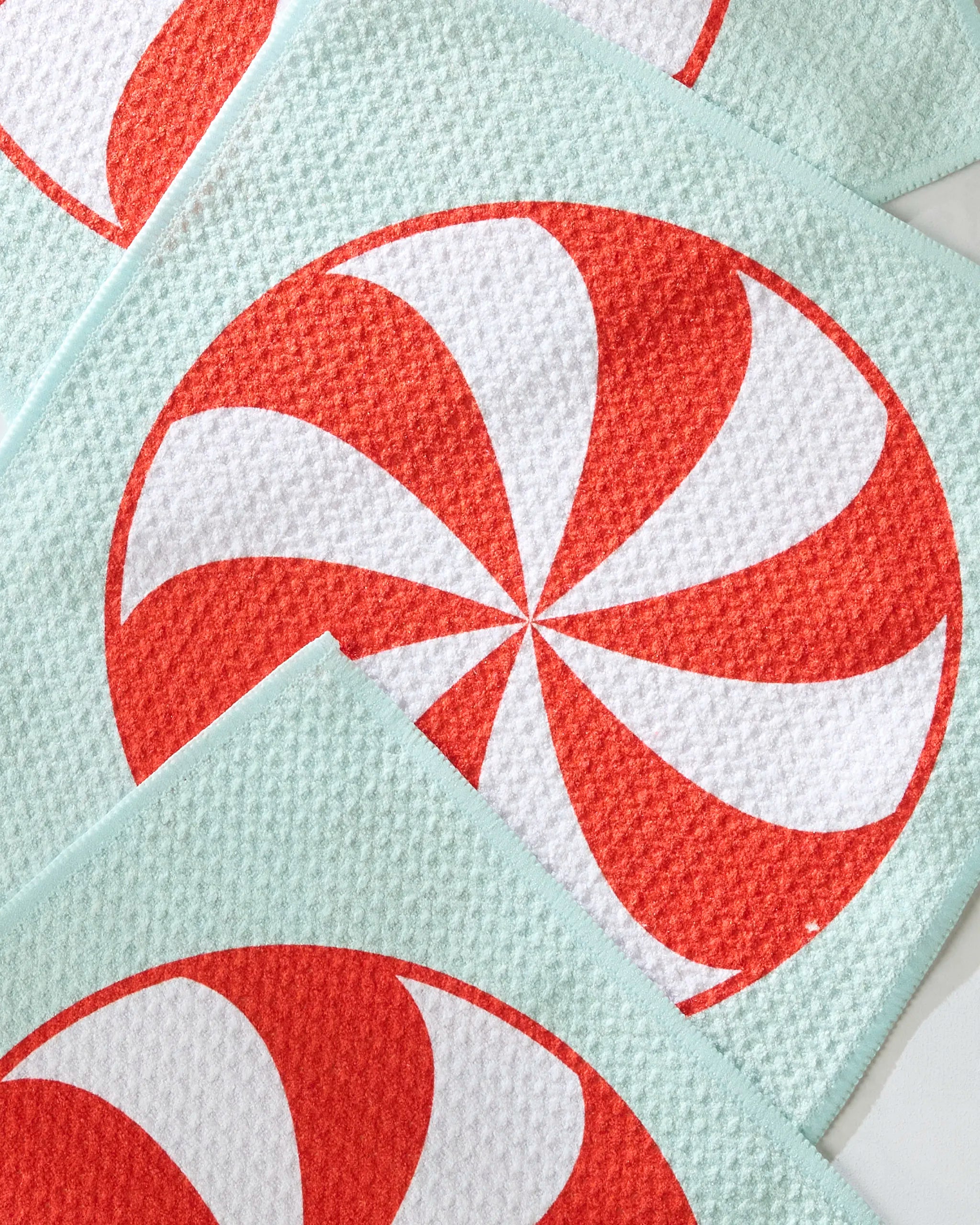 Geometry’s Peppermint Swirl dishcloths, made from recycled materials, feature a large red and white peppermint swirl design with teal borders and a textured white surface, shown overlapping in a flat lay.