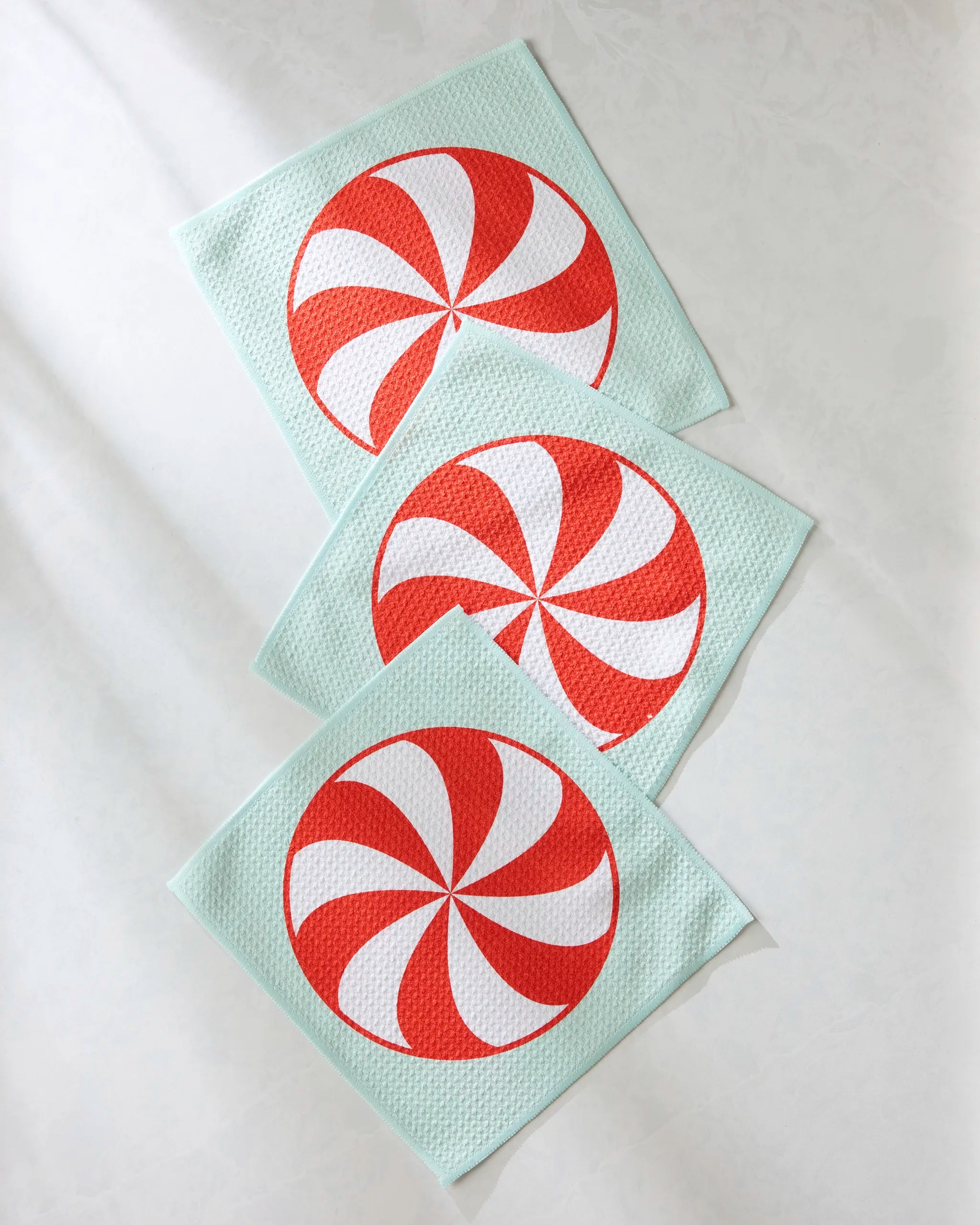 Three Peppermint Swirl dishtowels by Geometry, light blue with red and white swirl designs, are arranged diagonally on a light textured surface.