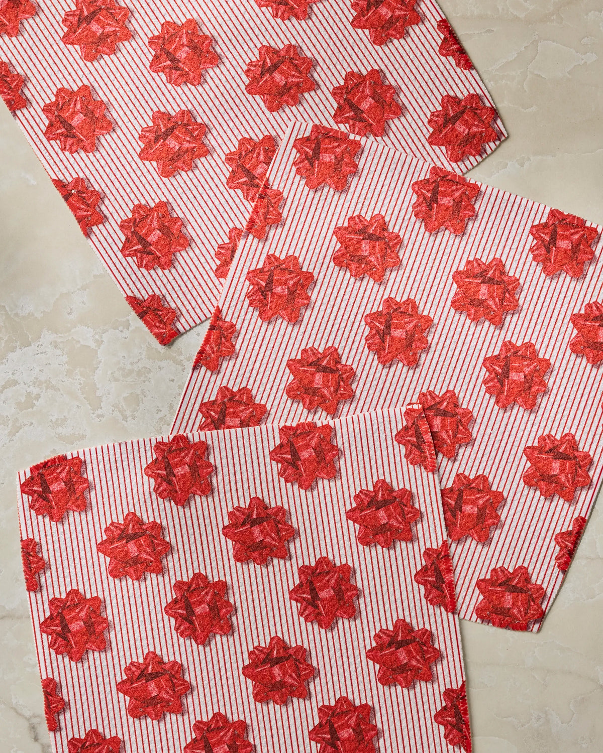 Three white “Perfectly Wrapped” gift bags from Geometry, featuring vertical red stripes and festive red bow patterns, are spread out on a light marble surface—creating an ideal backdrop for presenting a colorful Geometry dishcloth set or super-absorbent dishtowels made from recycled materials.