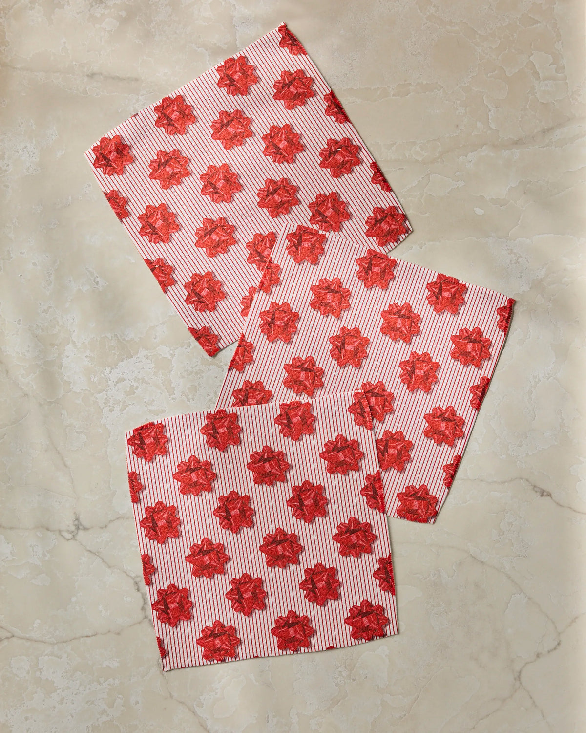 Three “Perfectly Wrapped” paper napkins by Geometry, featuring a red flower and stripe pattern and crafted from recycled materials, are casually arranged with slight overlap on a light beige marble surface.