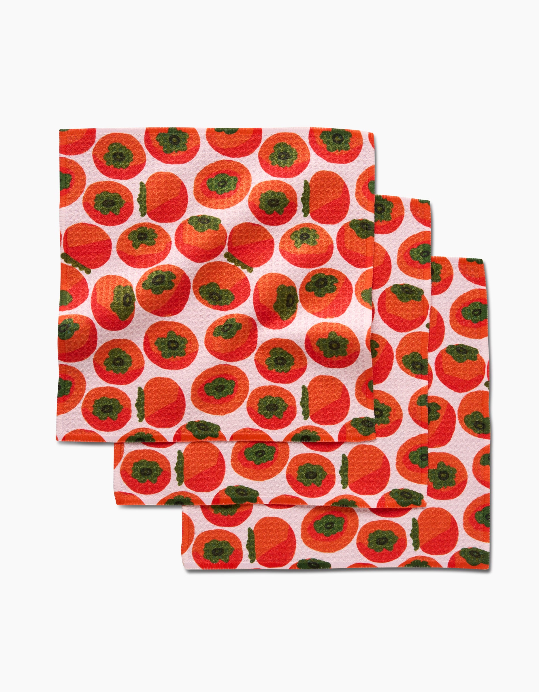 The Persimmons dishcloth set by Katie OShea features three folded Geoweave cloths with a stylish red-orange persimmon pattern and green tops on a white background, arranged in a staggered stack.