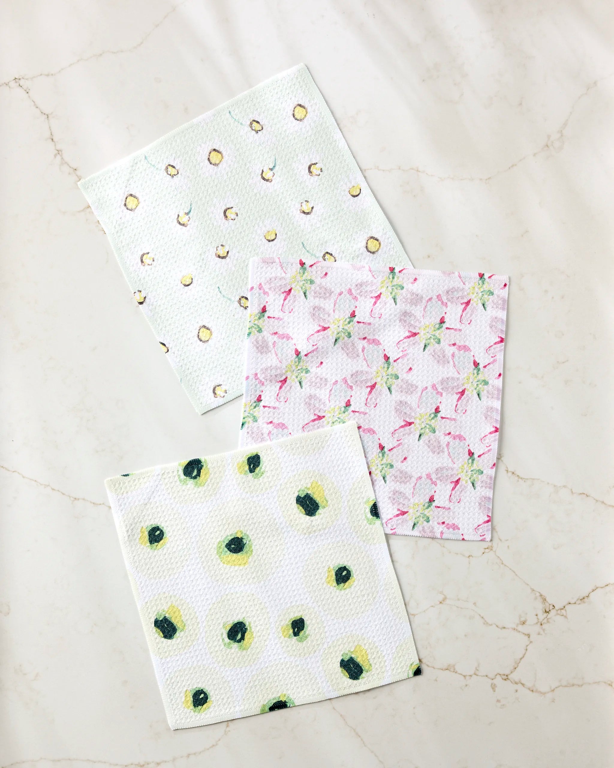 Three “Petals” paper napkins by Ania Zwara Art, made from recycled materials, are arranged on marble. The top has yellow and green dots, the middle shows pink flamingos, and the bottom is decorated with green avocados.
