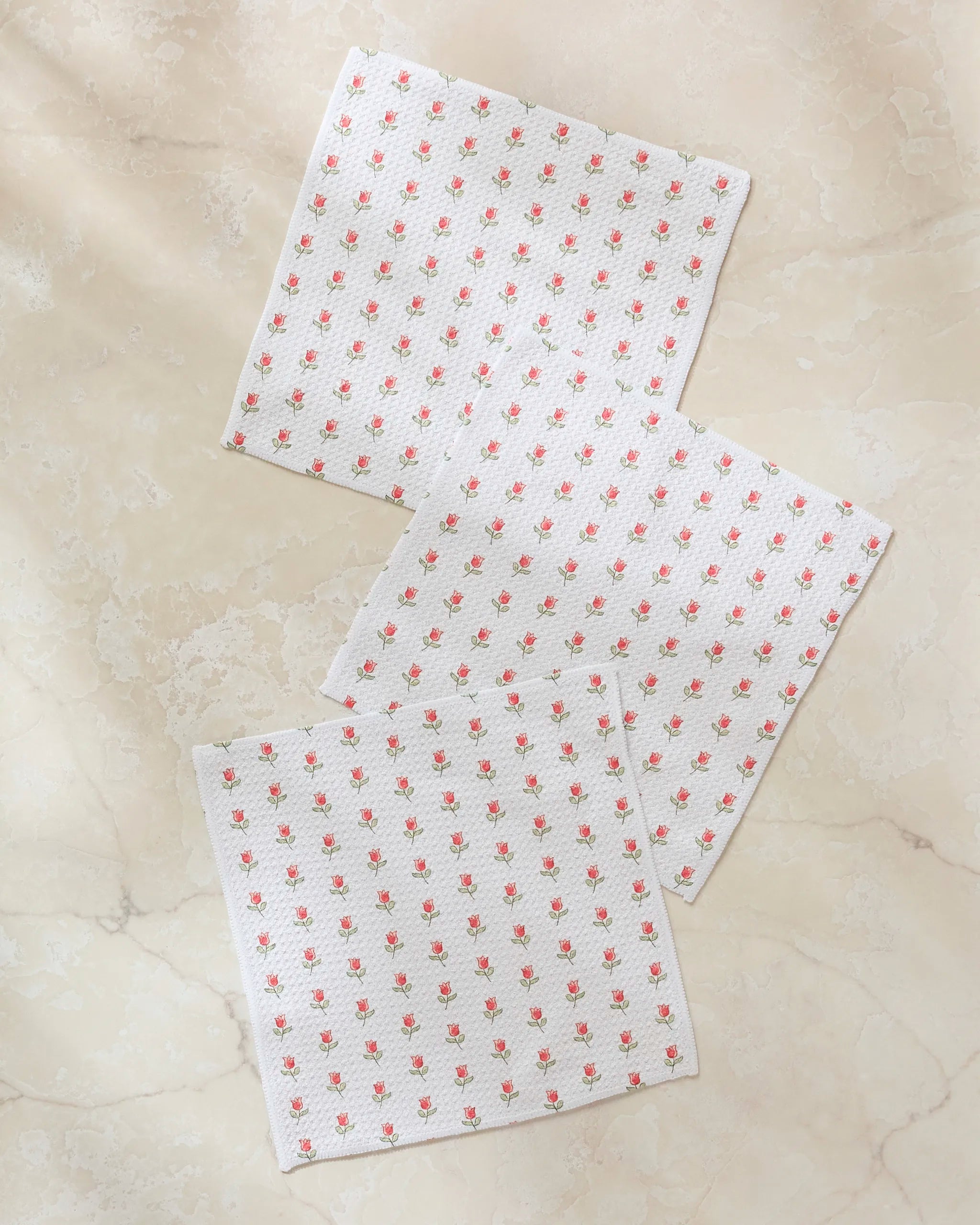 Three "Petite Tulip" kitchen towels by Danelys Sidron, featuring a small red floral pattern, are arranged in a scattered layout on a light beige marble surface.