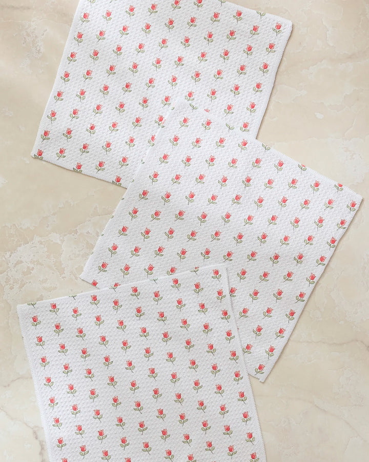 Four "Petite Tulip" kitchen towels by Danelys Sidron, featuring a pattern of small pink roses and green leaves on recycled fabric, are laid out slightly overlapping on a light beige marble surface.