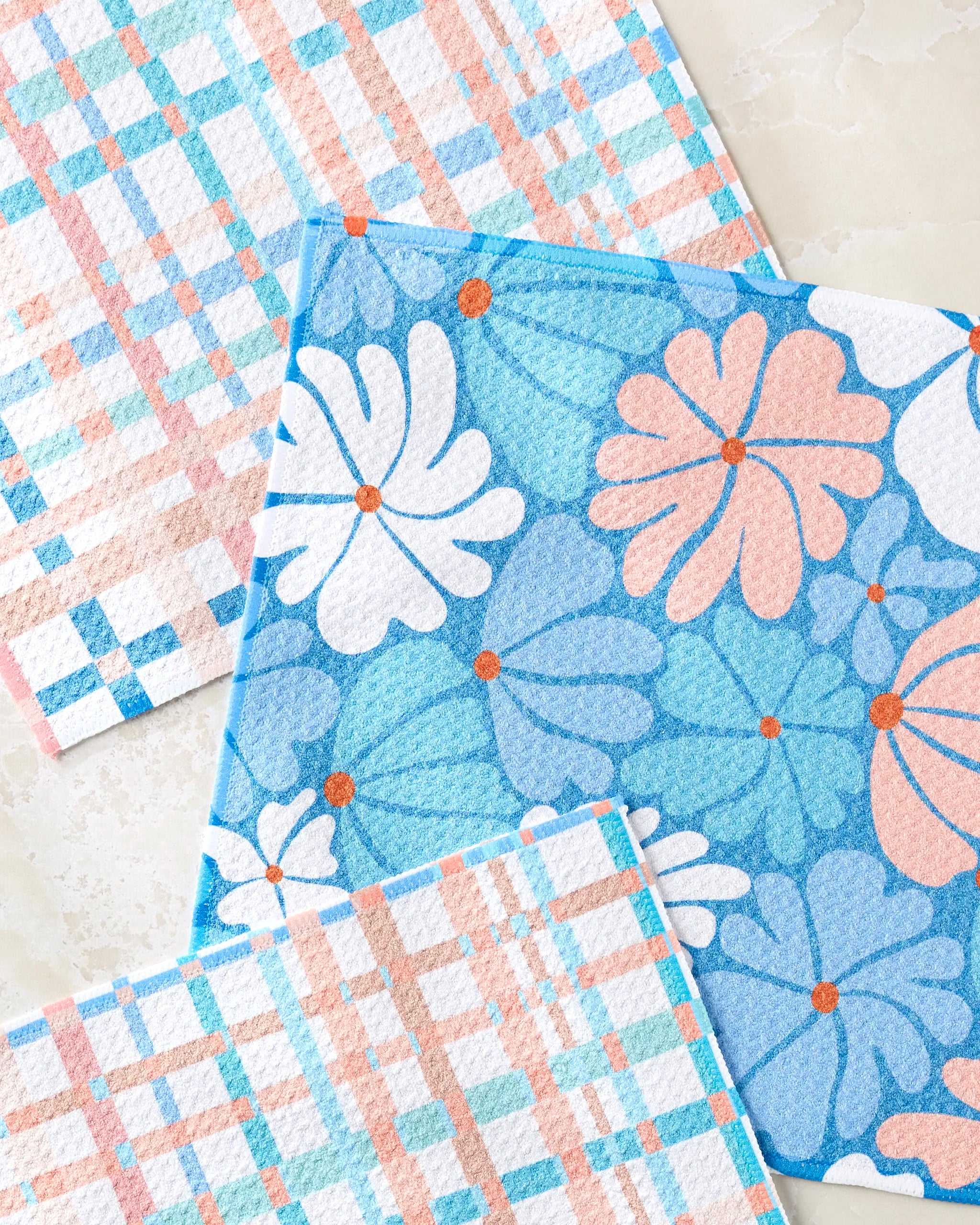The Rachel Parker "Picnic Flowers" set features three reusable cloths—two in blue, peach, and white plaid, and one with bold floral shapes—arranged vibrantly on a light, textured surface.