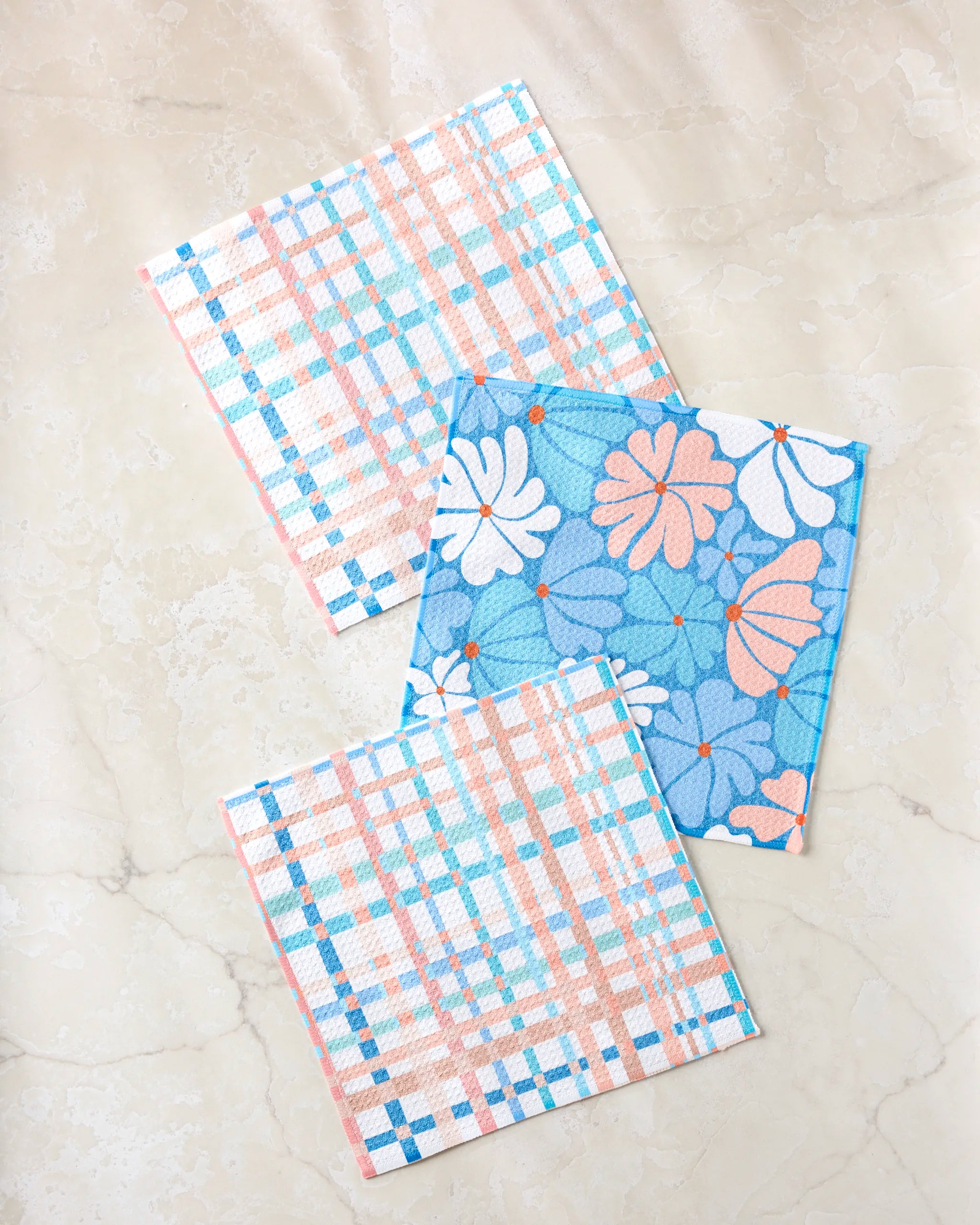 Three folded cloth napkins from the Picnic Flowers set by Rachel Parker are arranged on a light beige marble surface—two with a blue, pink, and white plaid pattern, and one with blue, orange, and white floral designs.