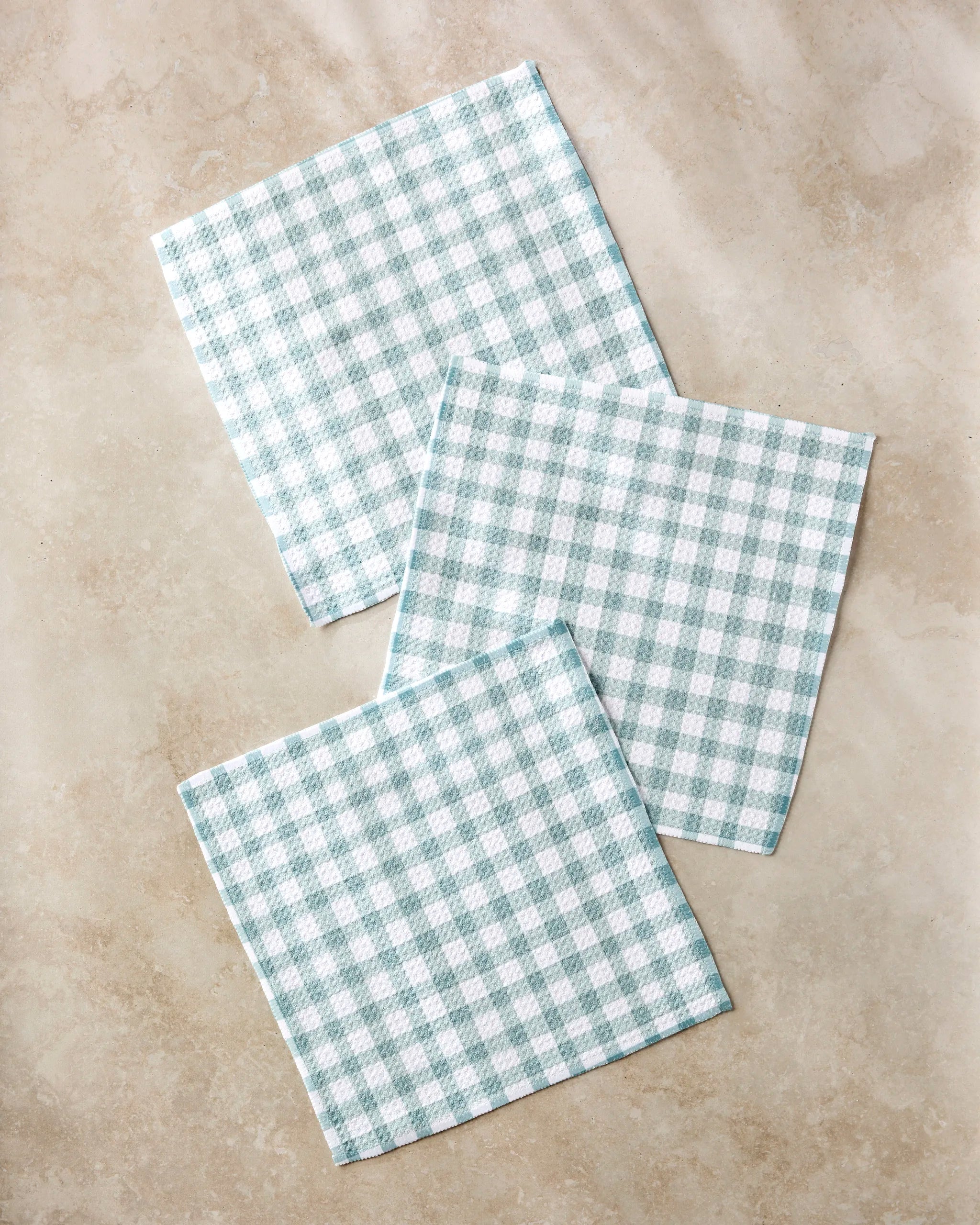 Three Picnic Gingham napkins by Hufton Studio, featuring blue and white checks, are laid out on a light beige stone surface with some overlap and slight angles between each napkin.