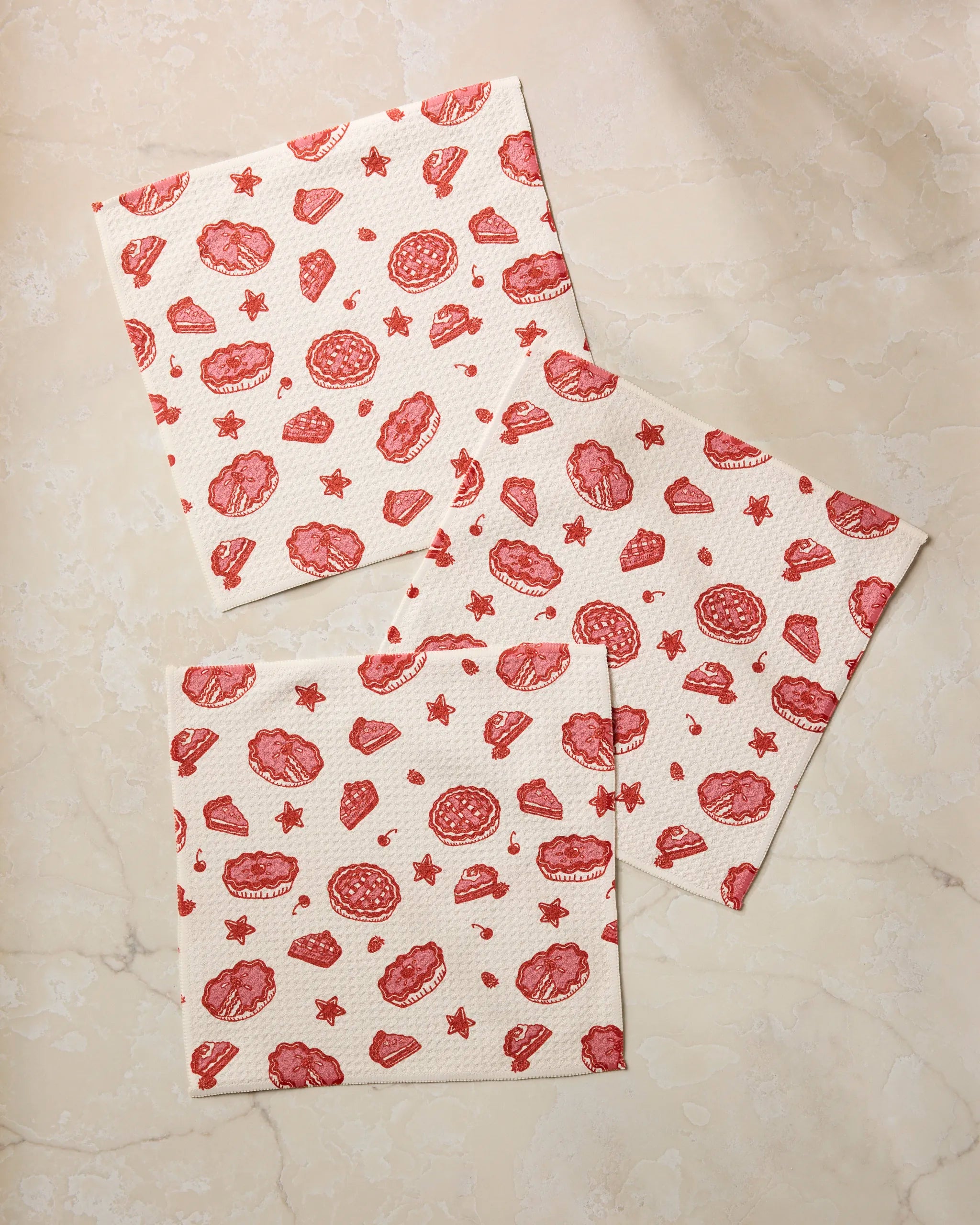 Three Pie-triotic square paper napkins by Geometry, featuring red cookie and star illustrations and made from recycled materials, are arranged on a light beige marble surface.