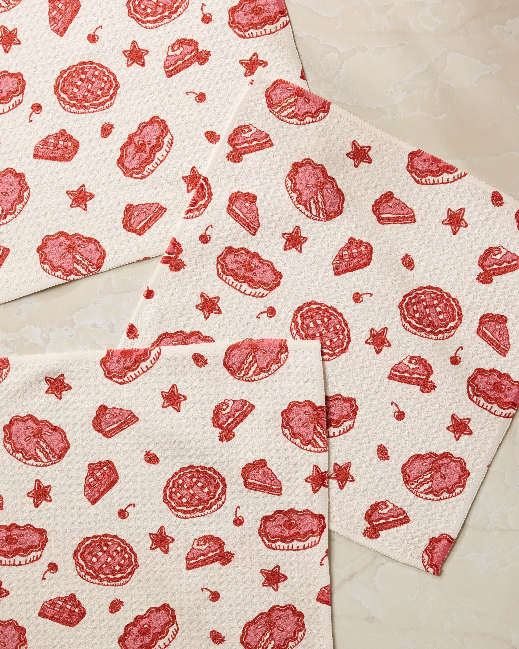 Three beige Pie-triotic napkins by Geometry, featuring red illustrations of pies, cake slices, and pastries, are arranged on a marble surface. This set is made from recycled materials and showcases a charming repeated dessert pattern.
