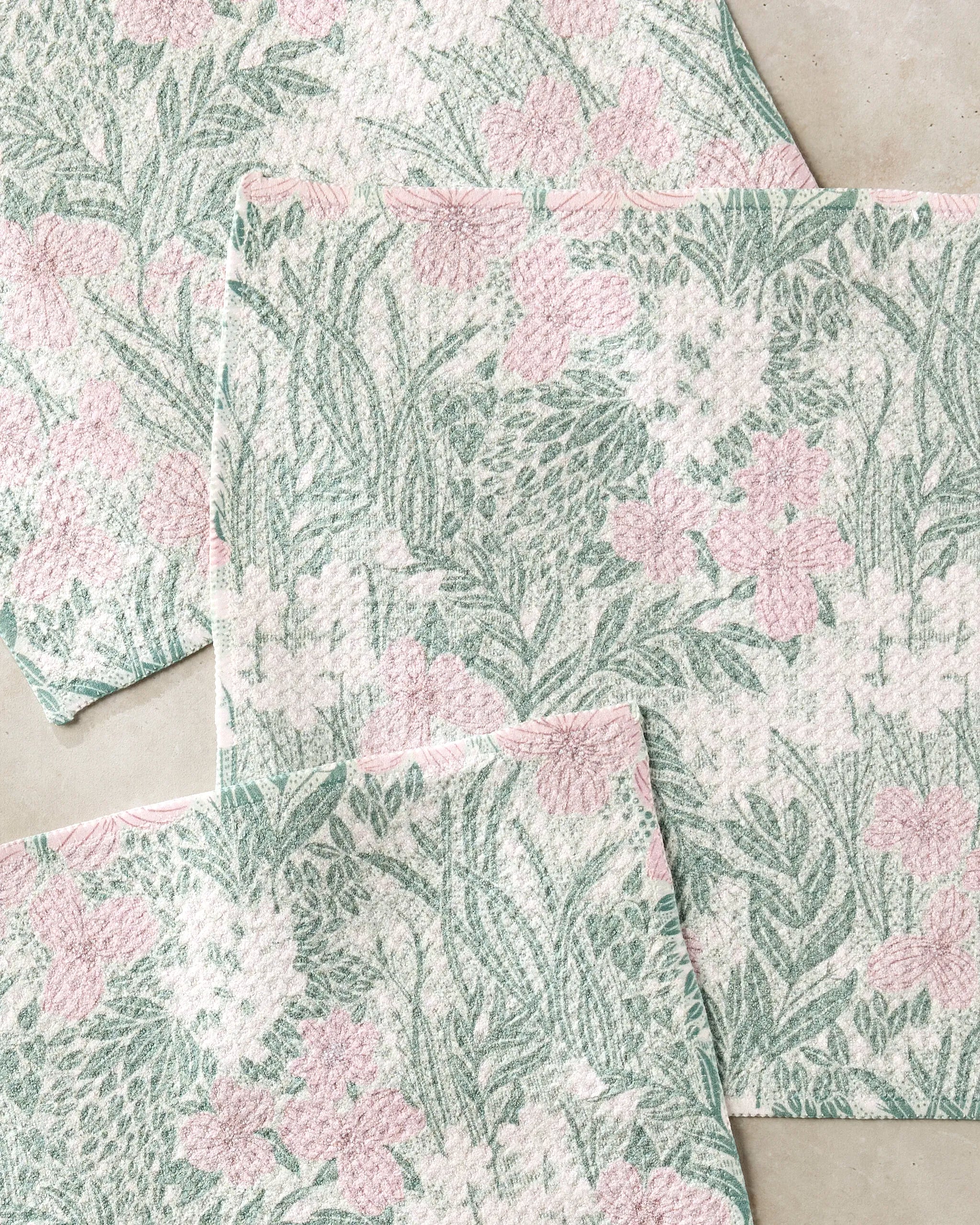 Four Pink and Sage Meadow cloths by Katie O’Shea, featuring overlapping pink and green botanical designs, rest on a light surface—an eco-friendly dishcloth set crafted from recycled materials.