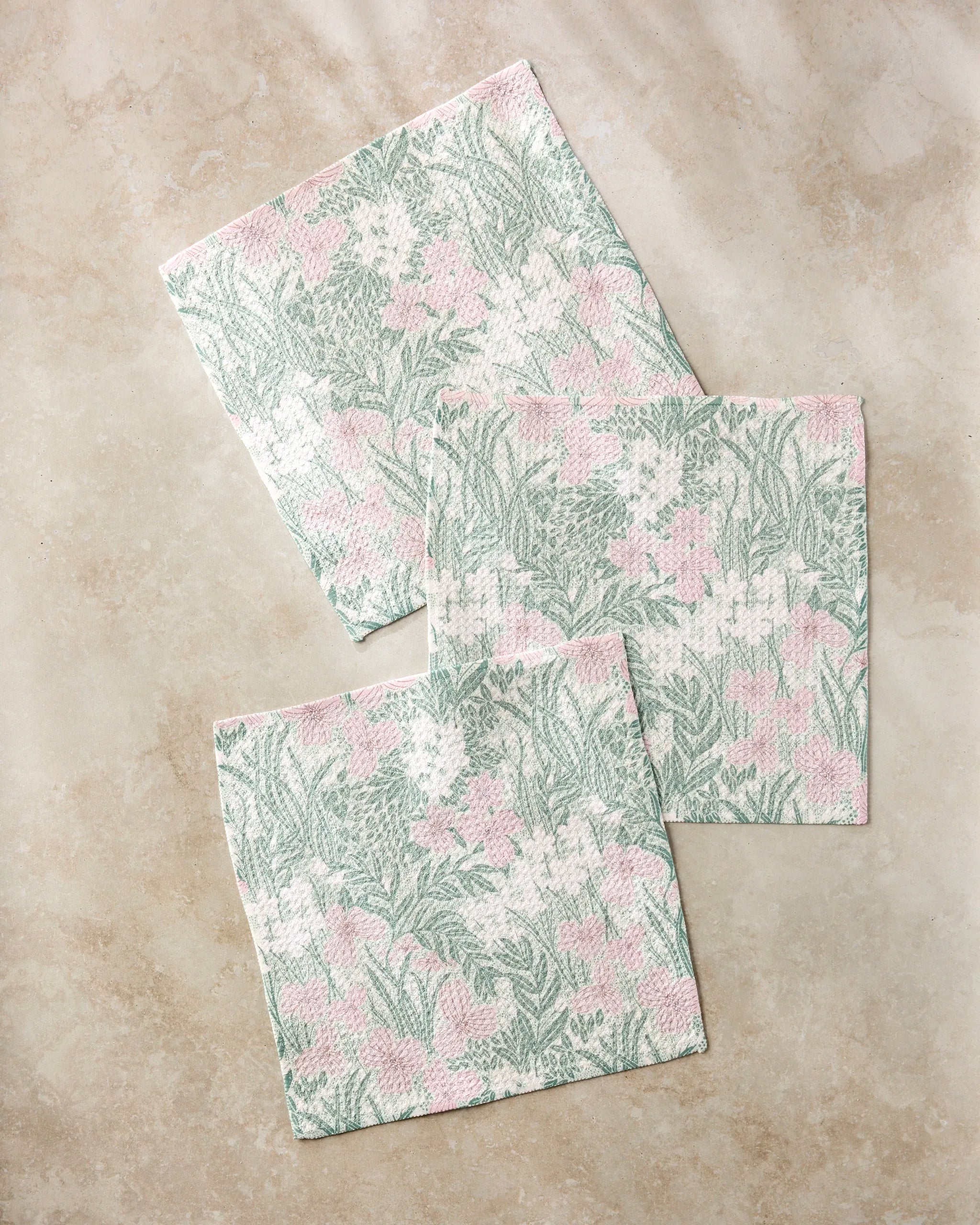 Three square napkins from Katie O'Shea's Pink and Sage Meadow collection, featuring soft green and pink florals, are arranged on a beige marble surface to create a charming kitchen dishcloth set.