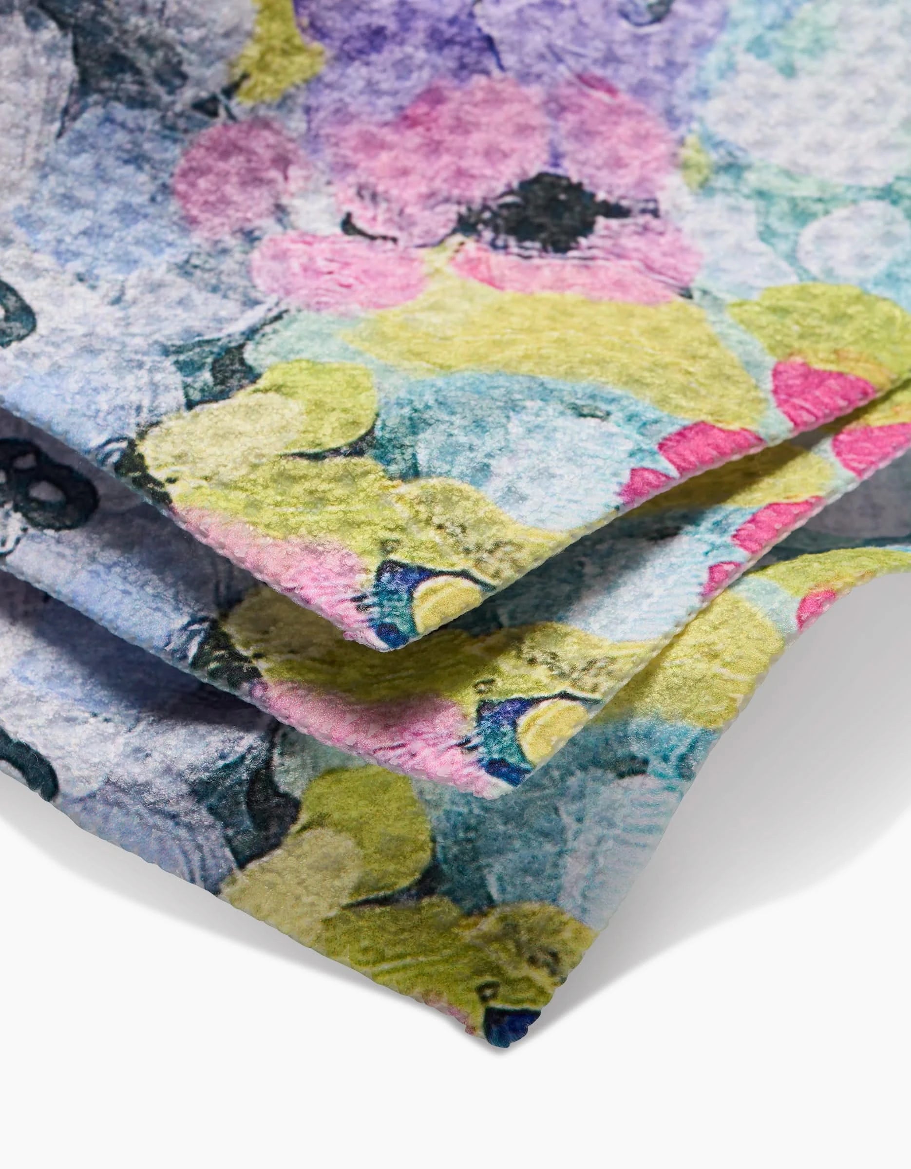 A close-up of the Pink Fields of Joy Geoweave dishcloth by Ania Zwara Art, showing its textured surface and watercolor floral print in purple, pink, yellow, green, and blue tones. Soft shadows highlight the fabric’s intricate design.