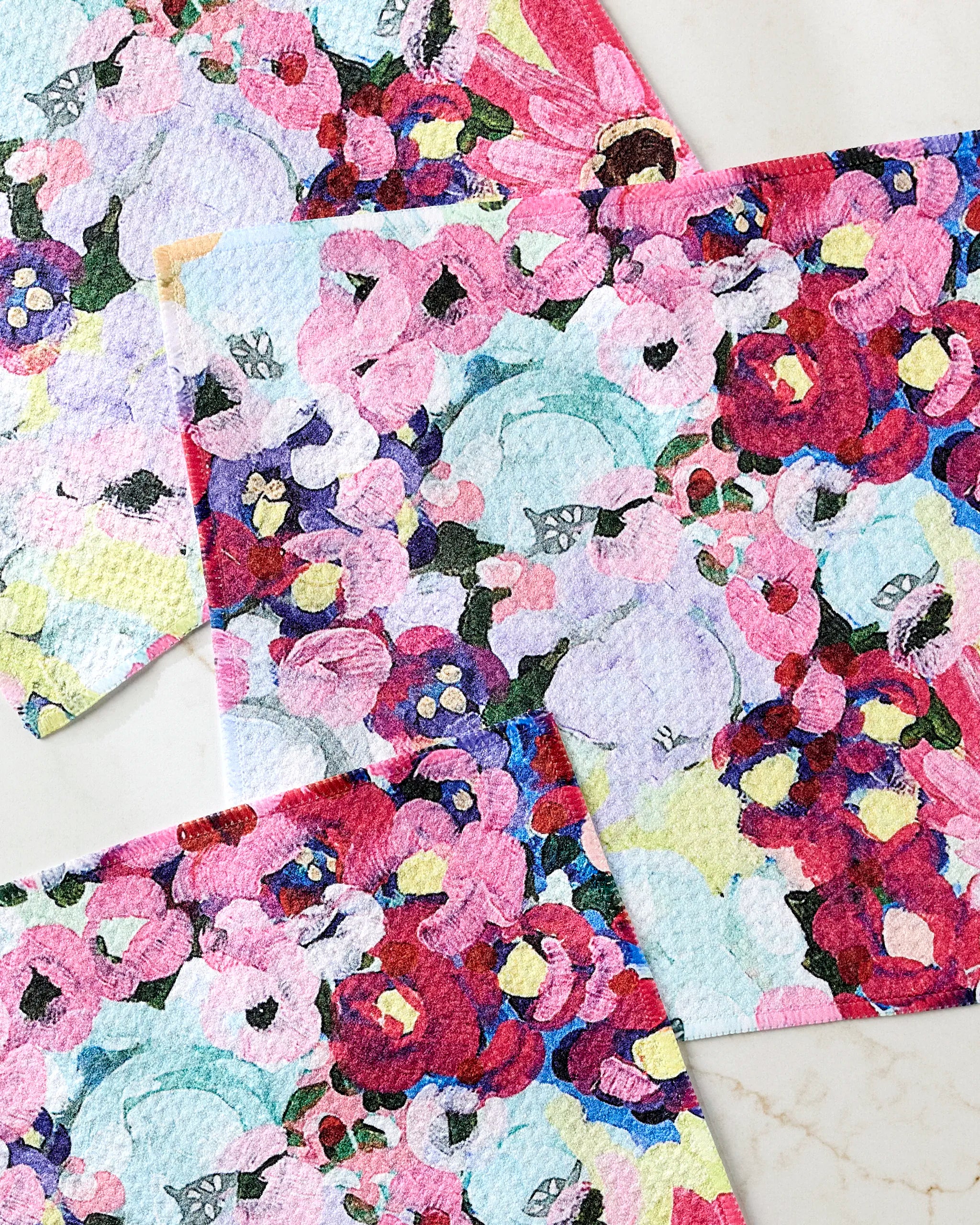 The Pink Fields of Joy dishcloth set by Ania Zwara Art features three rectangular cloths with a vibrant abstract floral pattern in pink, red, purple, blue, yellow, and white—made from recycled materials and arranged overlapping on a light surface.