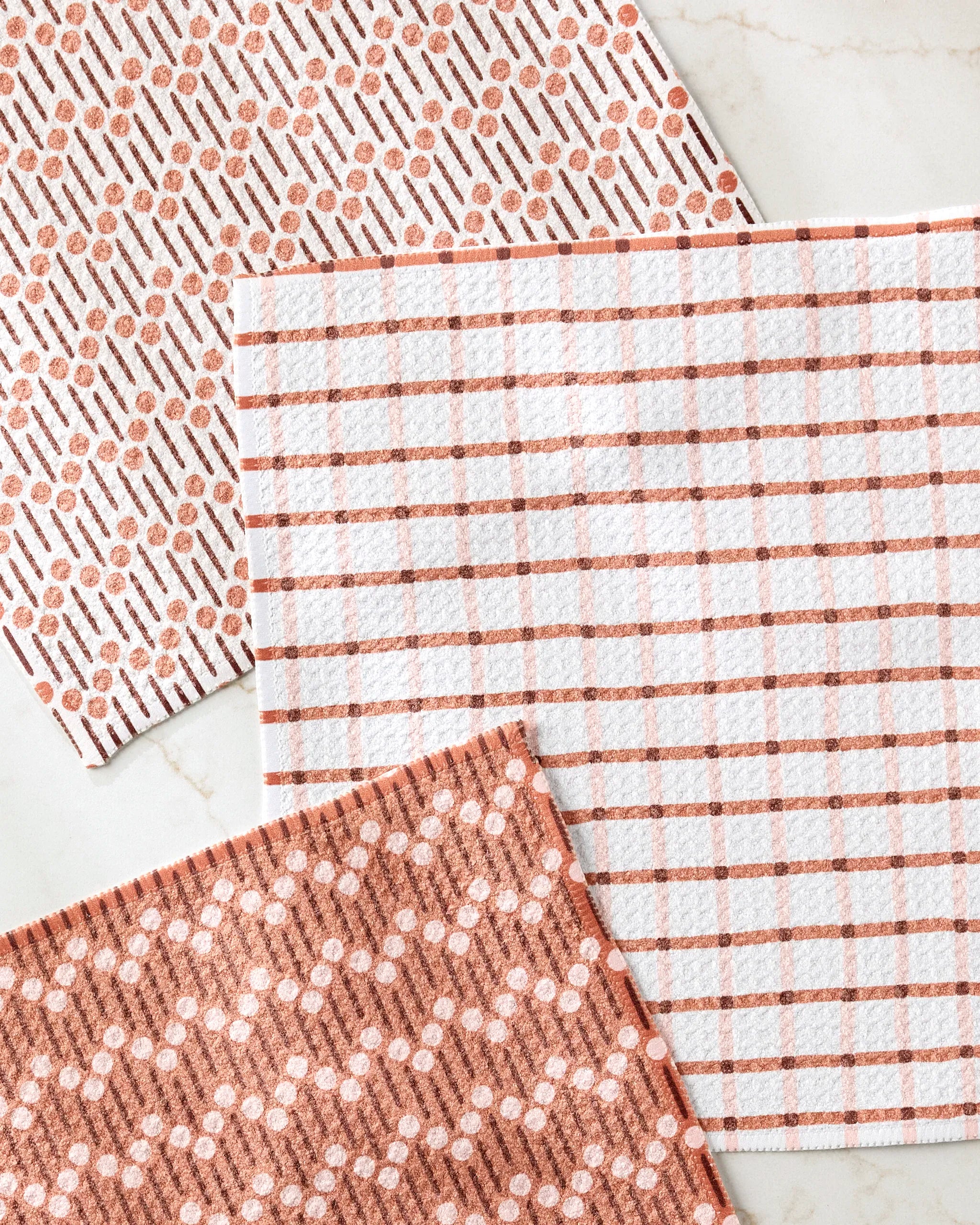 Three "Playful Retro" dish towels by Teri the Butter are arranged on a light marble surface. Each features a unique terracotta and white geometric pattern: dots and dashes, grid lines, and intersecting circles with lines.