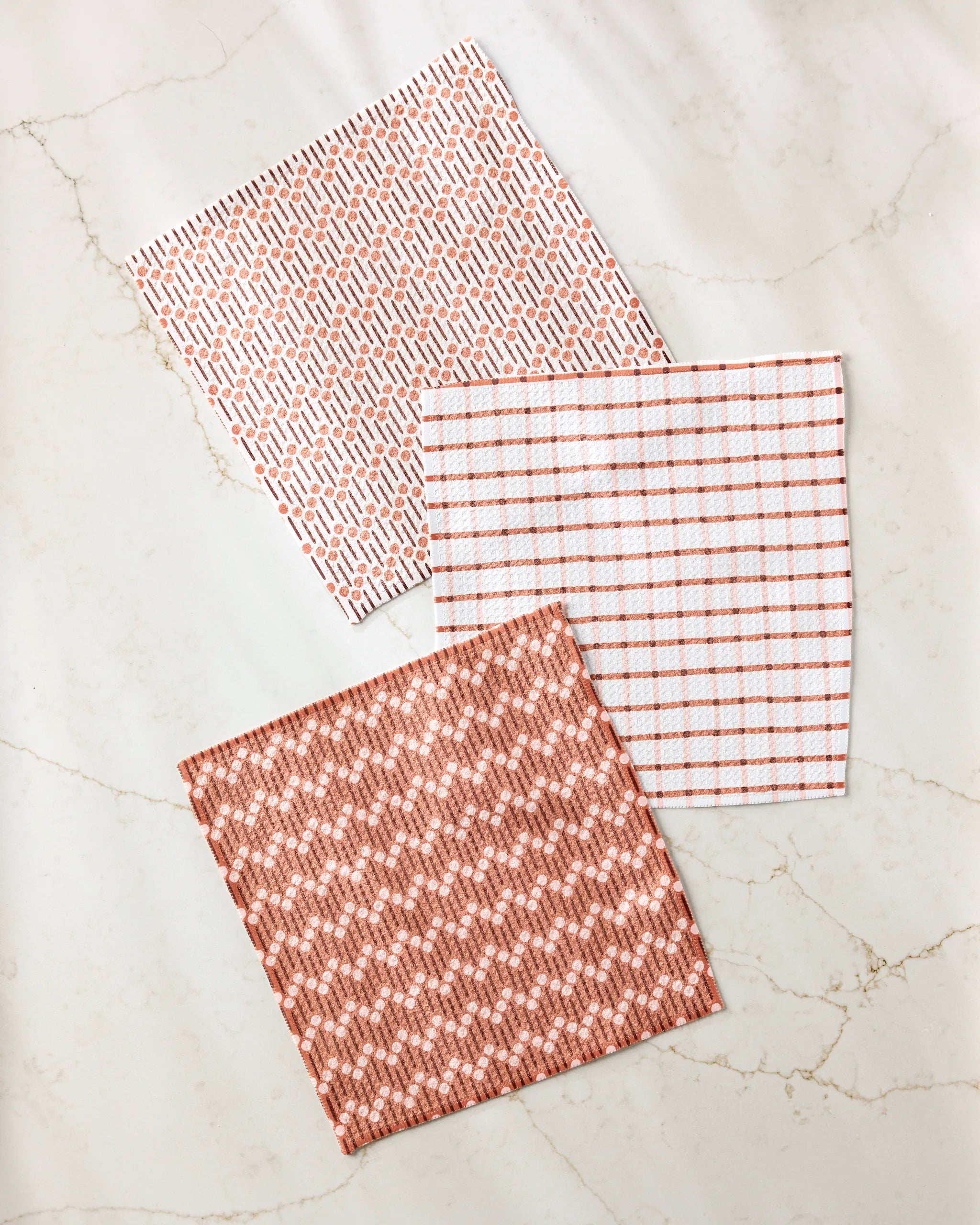 Three Playful Retro square cloth napkins by Teri the Butter—made from recycled materials with red and white dash, grid, and woven patterns—are displayed on a light marble surface.