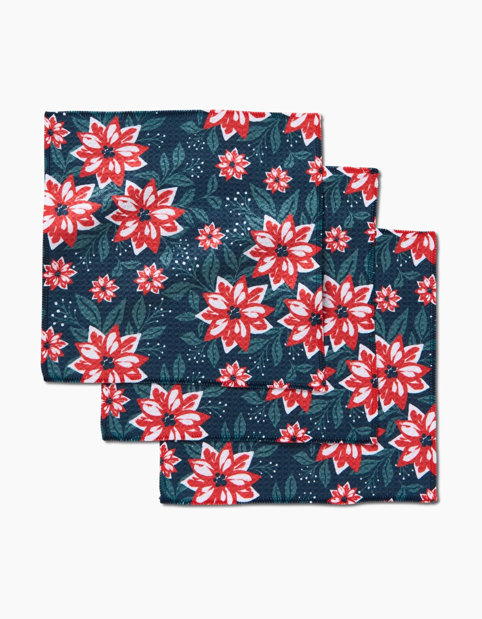 Three Poinsettia Garden napkins by Charlie Rowan Designs, made from Geoweave fabric, display a red and white floral pattern with green leaves on dark blue and are folded and stacked against a white background.