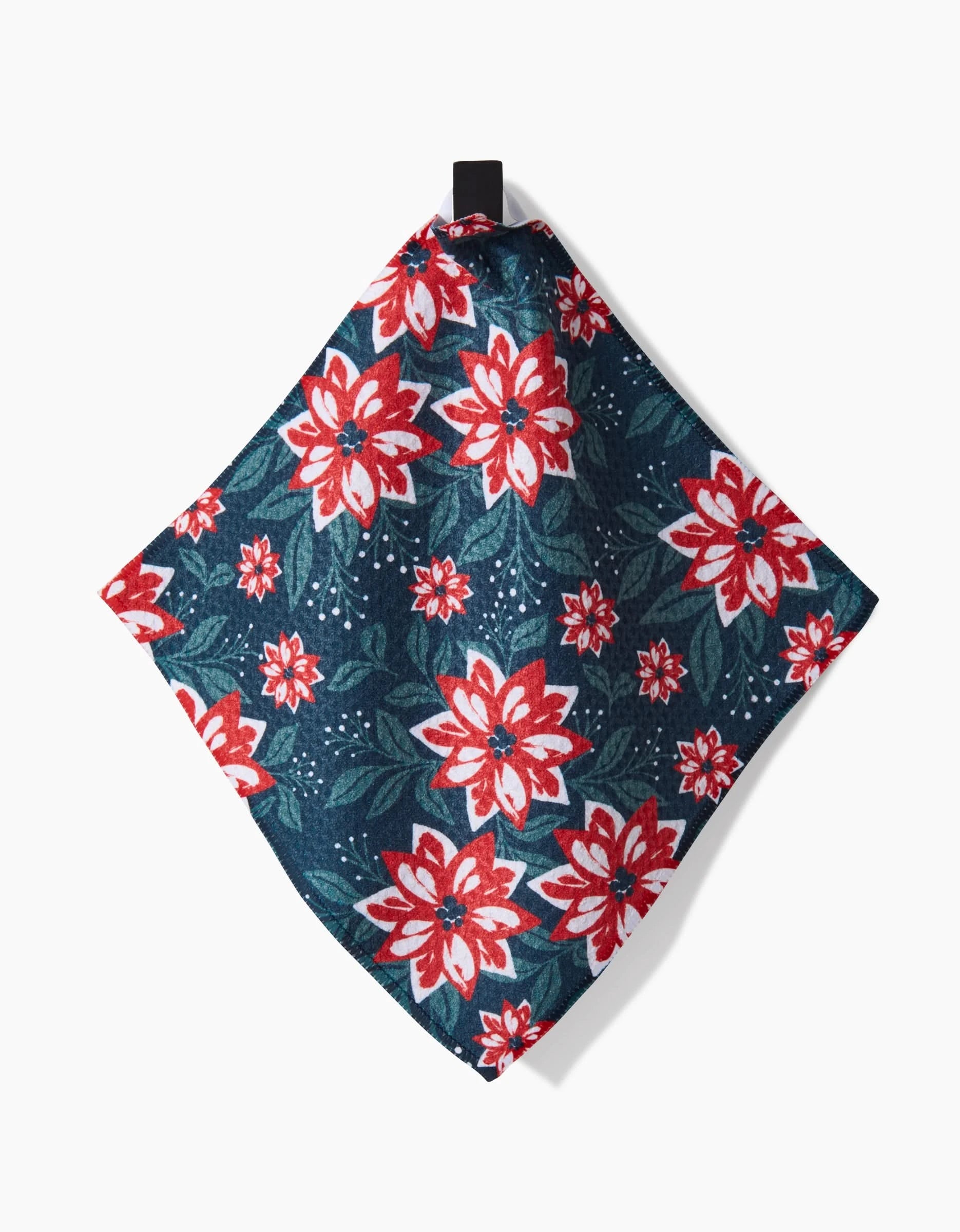 The Charlie Rowan Designs Poinsettia Garden kitchen towel, made from Geoweave fabric, features red and white poinsettias with green leaves on a dark blue background. It hangs from a black loop against a white backdrop.