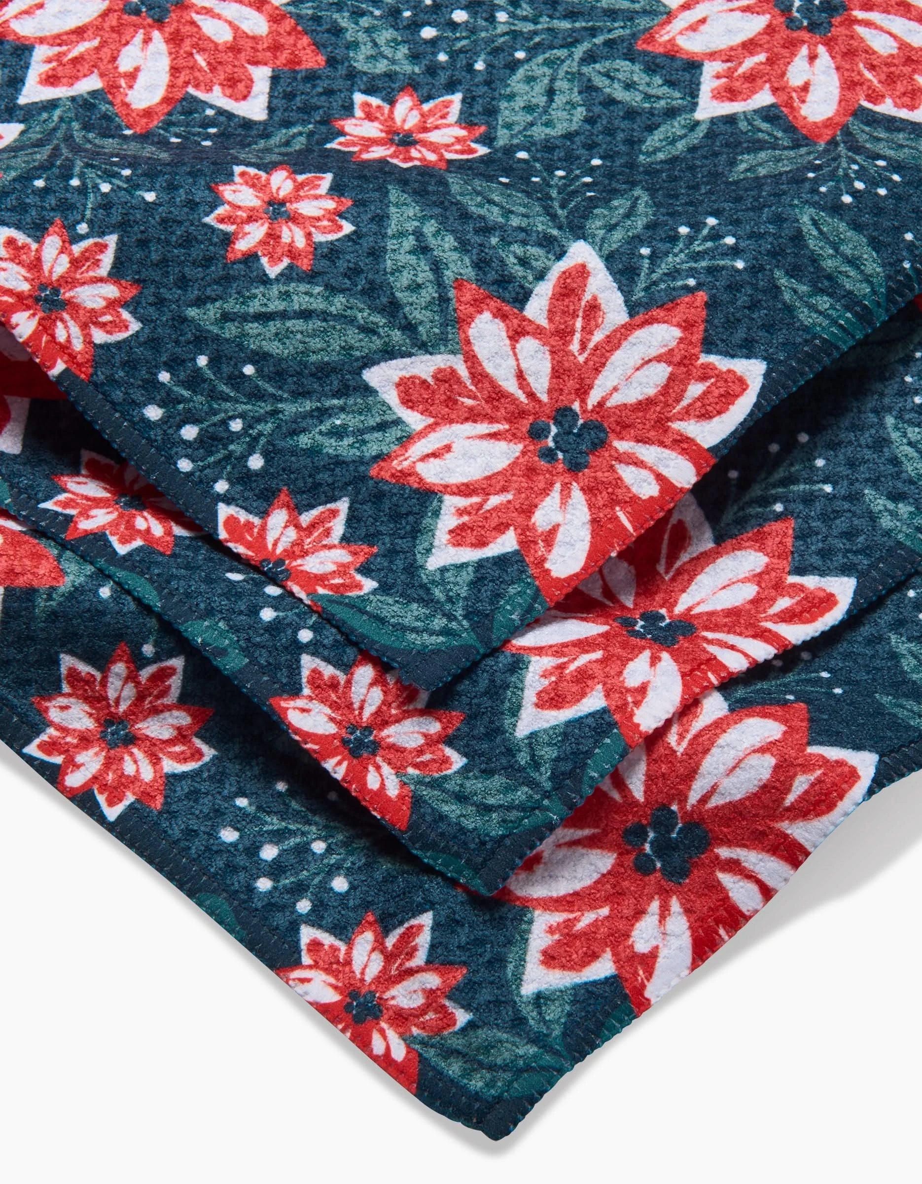 A close-up of the Poinsettia Garden kitchen towel by Charlie Rowan Designs, showcasing a vibrant floral pattern of red and white flowers with green leaves and white accents on a dark blue Geoweave fabric.