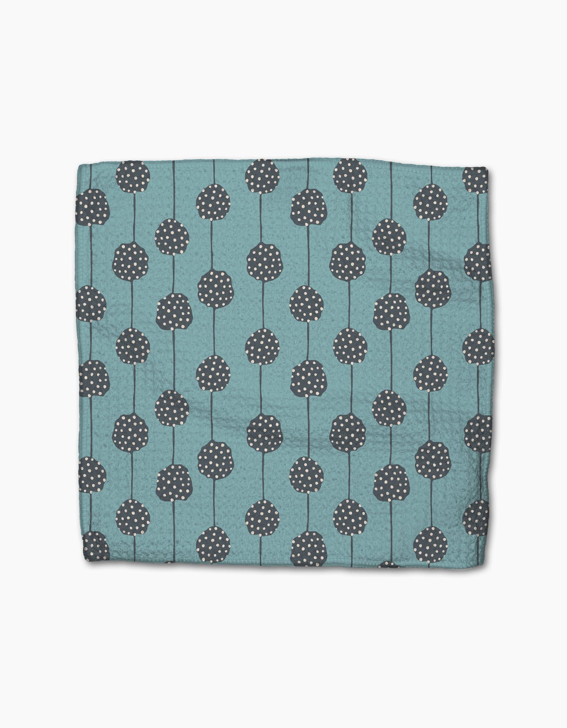 The Polka Dot Blooms napkin by Lizzie Clark is a folded teal cloth made from recycled materials, featuring dark gray circular patterns with white dots and vertical lines, displayed on a plain white background.