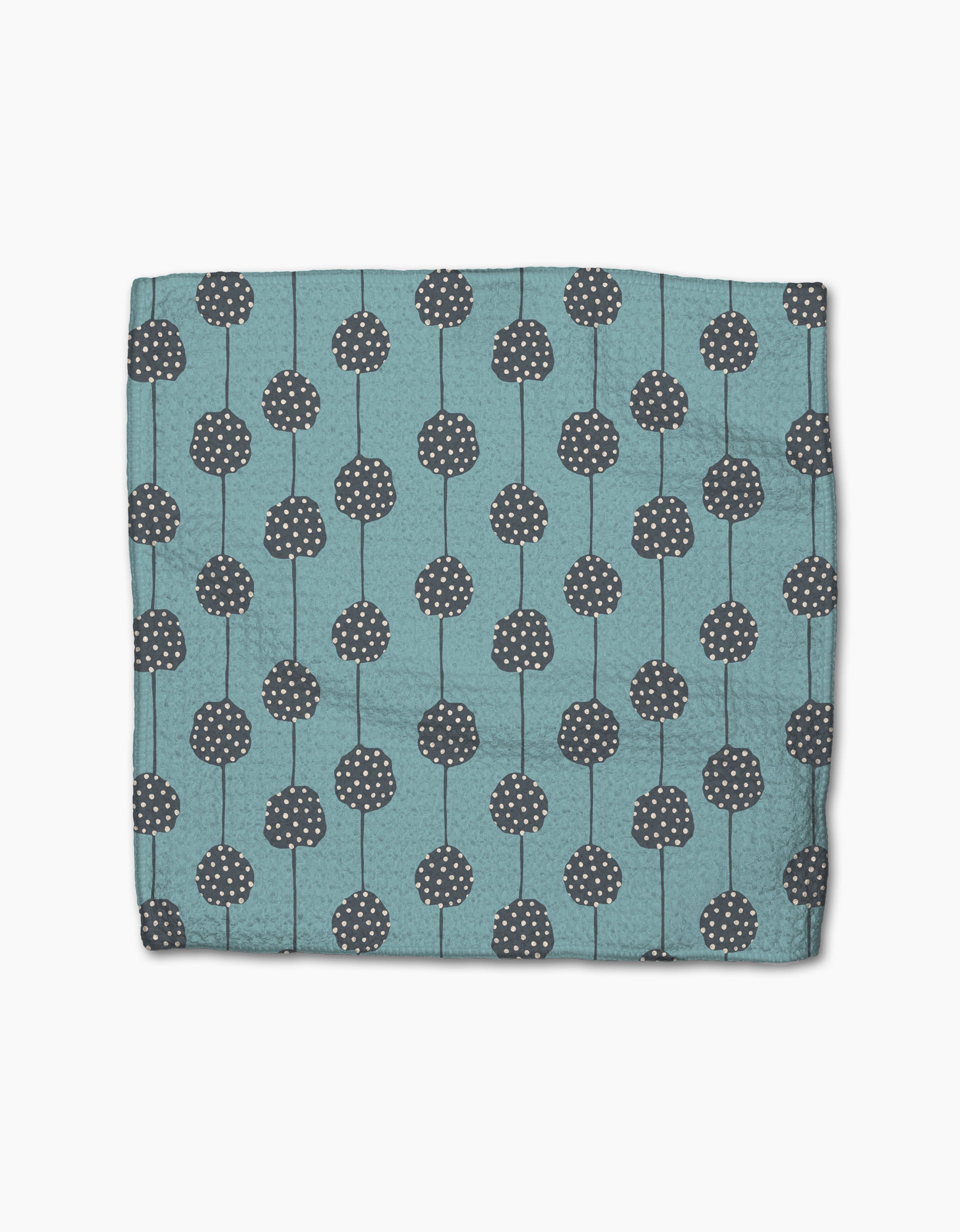 The Polka Dot Blooms napkin by Lizzie Clark is a folded teal cloth made from recycled materials, featuring dark gray circular patterns with white dots and vertical lines, displayed on a plain white background.