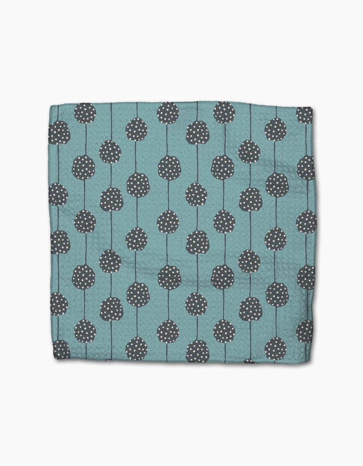 The Polka Dot Blooms napkin by Lizzie Clark is a folded teal cloth made from recycled materials, featuring dark gray circular patterns with white dots and vertical lines, displayed on a plain white background.