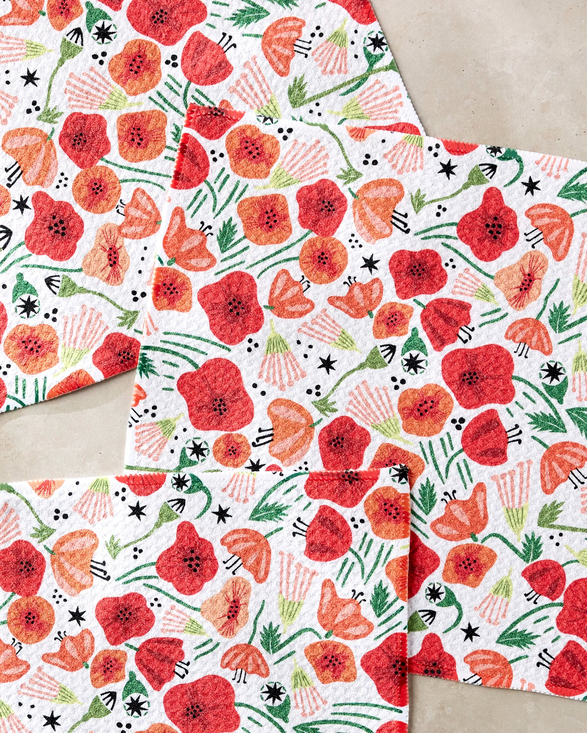 Three absorbent Elisabeth Wing "Poppy Power" dishcloths—featuring red and orange flowers, green leaves, and black and pink accents—overlap on a neutral surface, creating a stylish floral set for your kitchen.