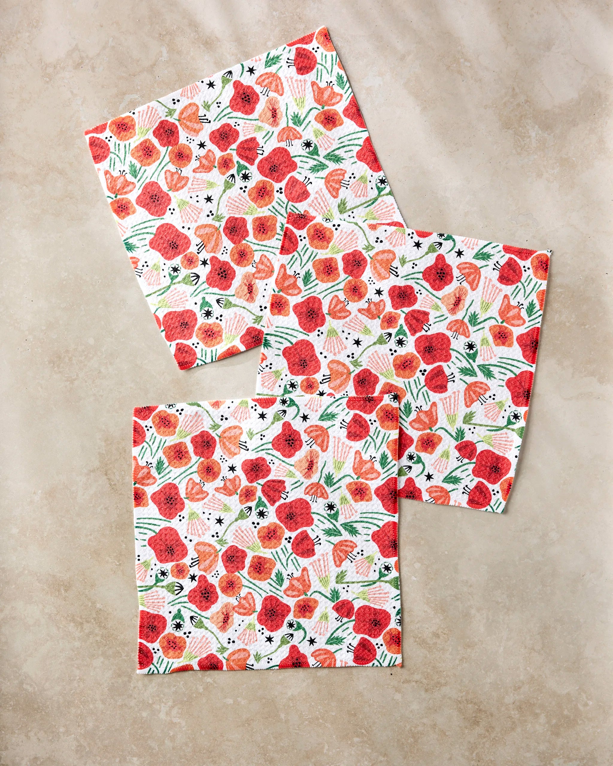 Three square pieces from Elisabeth Wing’s “Poppy Power” absorbent dishcloth set, featuring a red and orange floral pattern with green leaves and black accents, are arranged on a light beige surface.