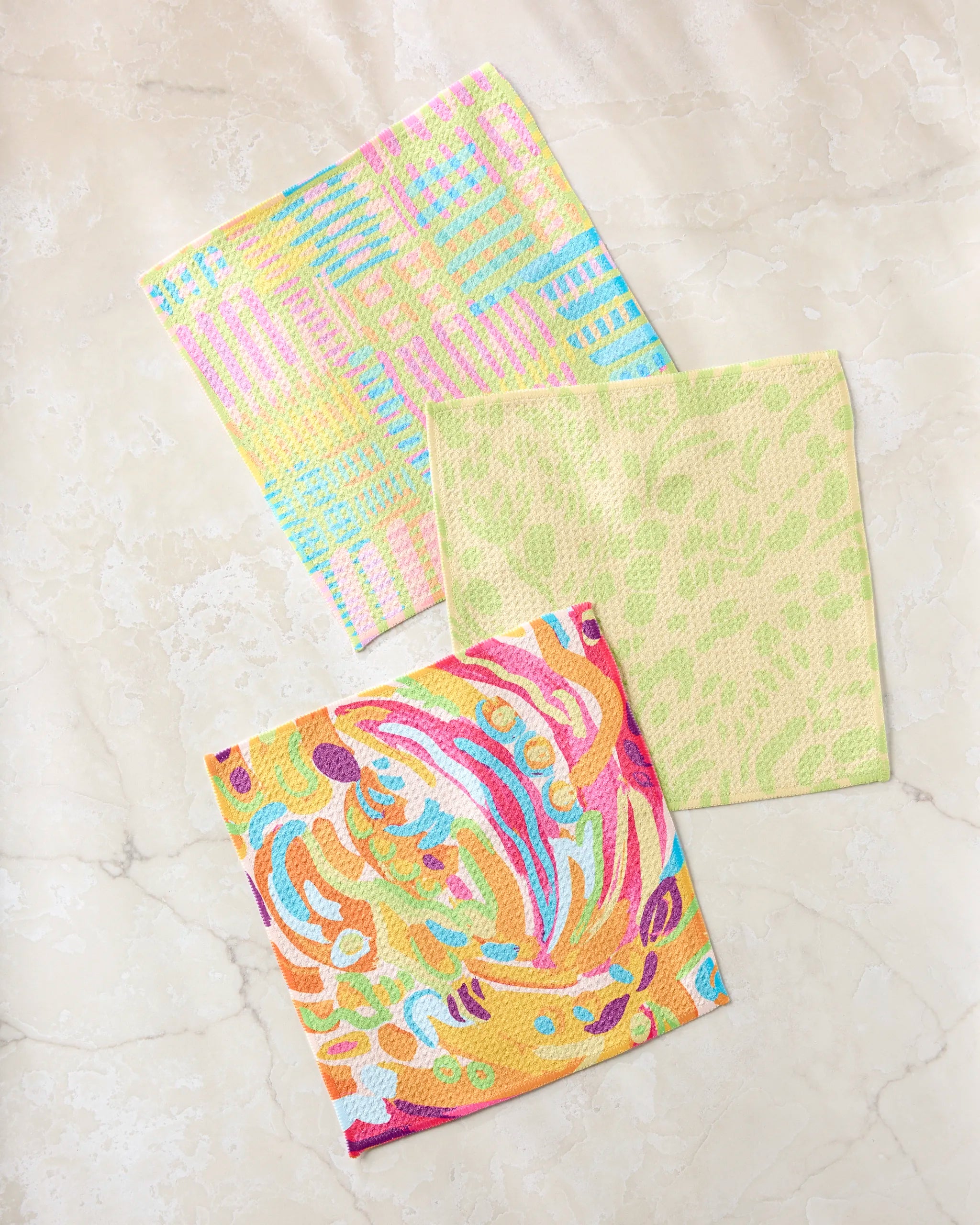 The Positive Vibes sponge cloth set by Sareka Unique includes three eco-friendly dishcloths with plaid, abstract swirls, and leaf prints, made from recycled materials and displayed on a light beige marble surface.