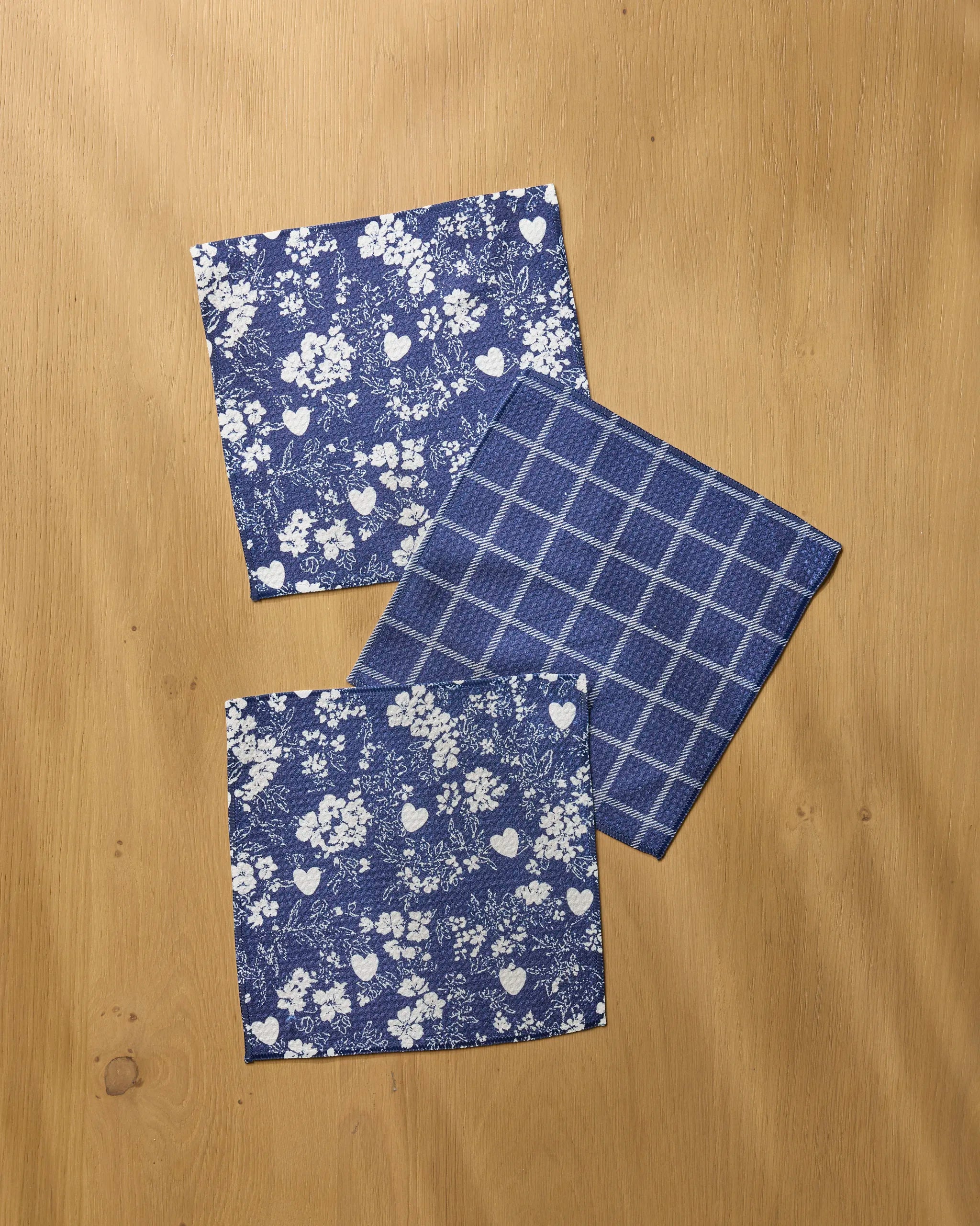 Three blue Posy Plaid fabric napkins from Geometry are arranged on light wood—two with a white floral and heart pattern and one in blue check, making them ideal absorbent dishcloths for any kitchen.