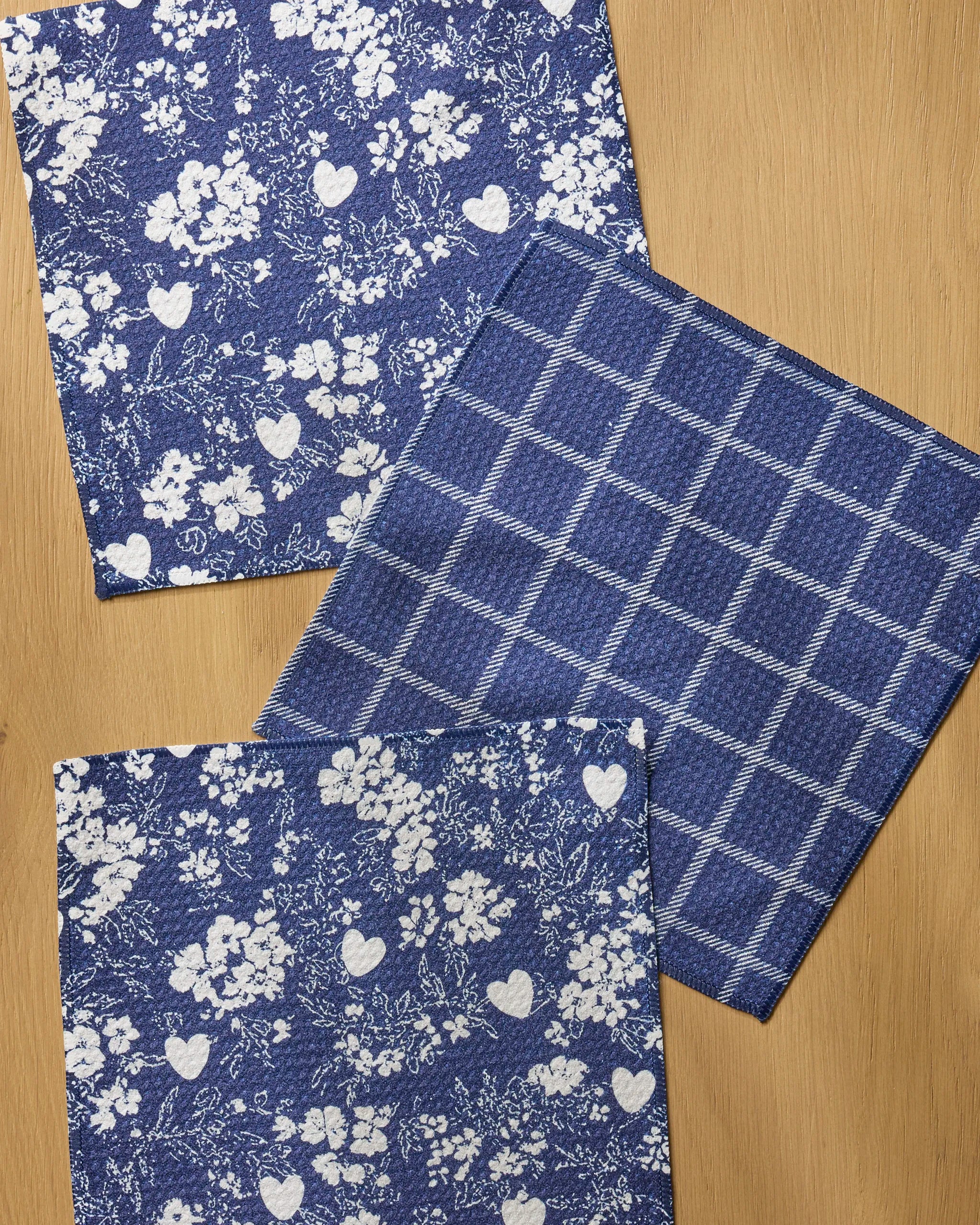 Three Posy Plaid cloth napkins by Geometry are displayed on wood. Two feature blue and white floral and heart patterns, while the third in this absorbent dishcloth set shows a checkered grid design.