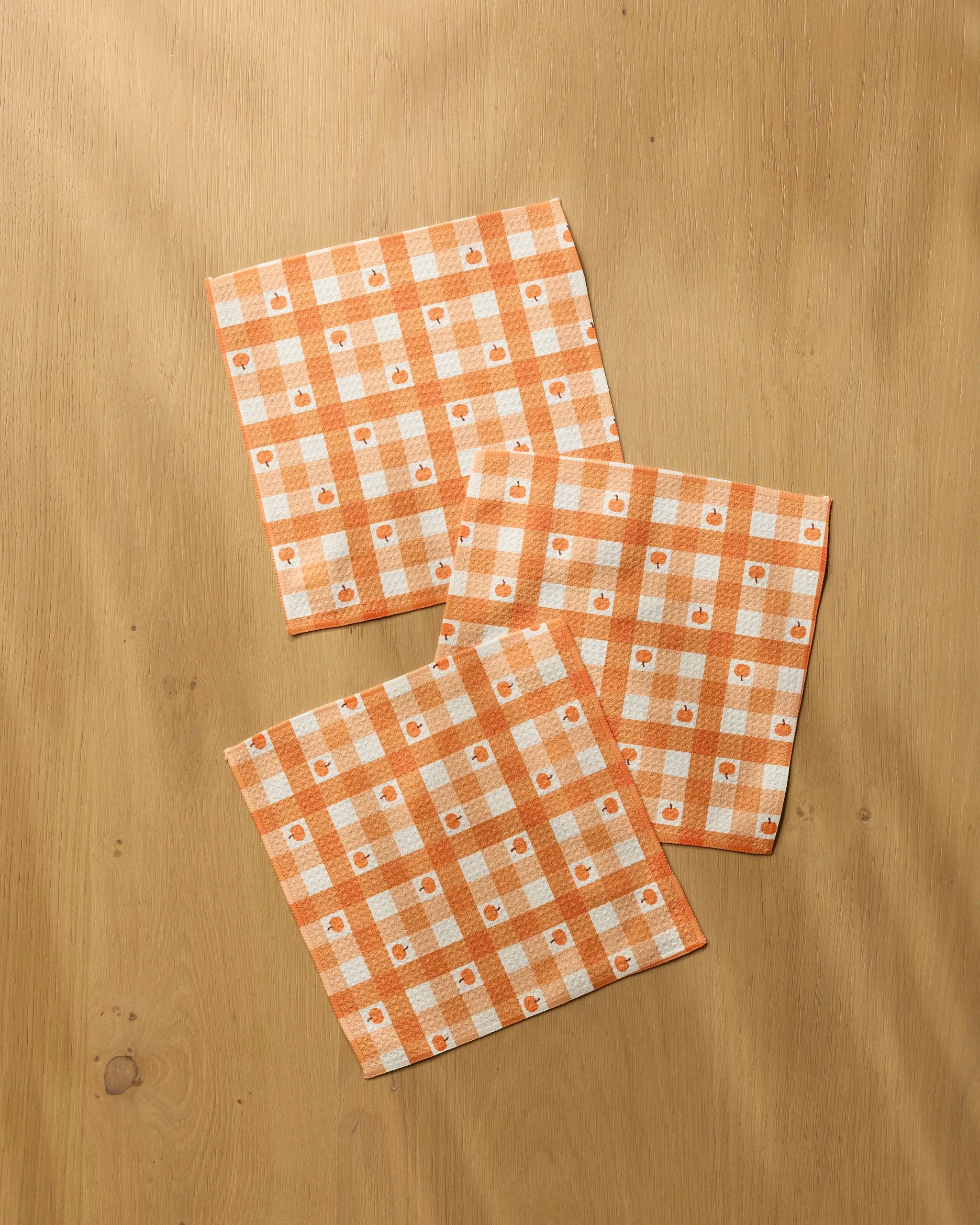 Three "Pumpkin Check" cloth napkins by Geometry, featuring an orange and white check pattern and small apples, made from recycled materials, are displayed on a light wood surface.