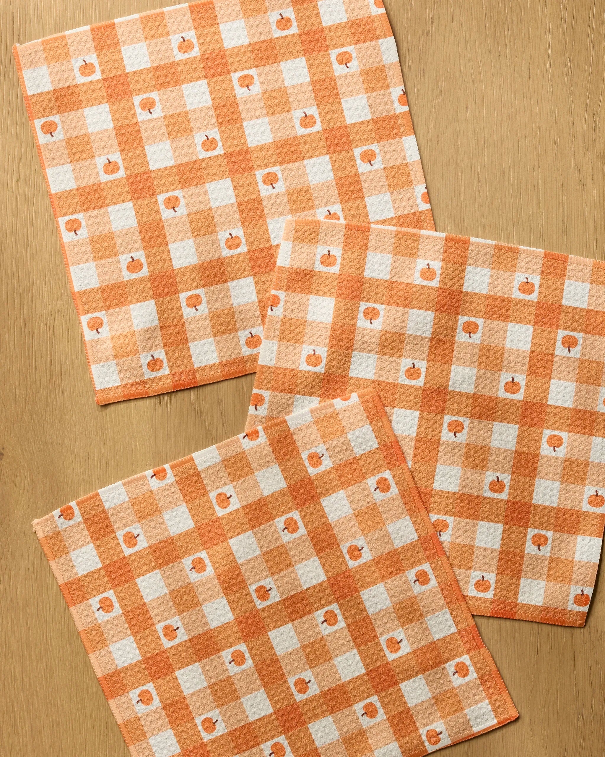 The Pumpkin Check dish towels by Geometry, featuring orange and white checkered patterns with small pumpkins, are made from recycled materials and displayed on a light wooden surface.