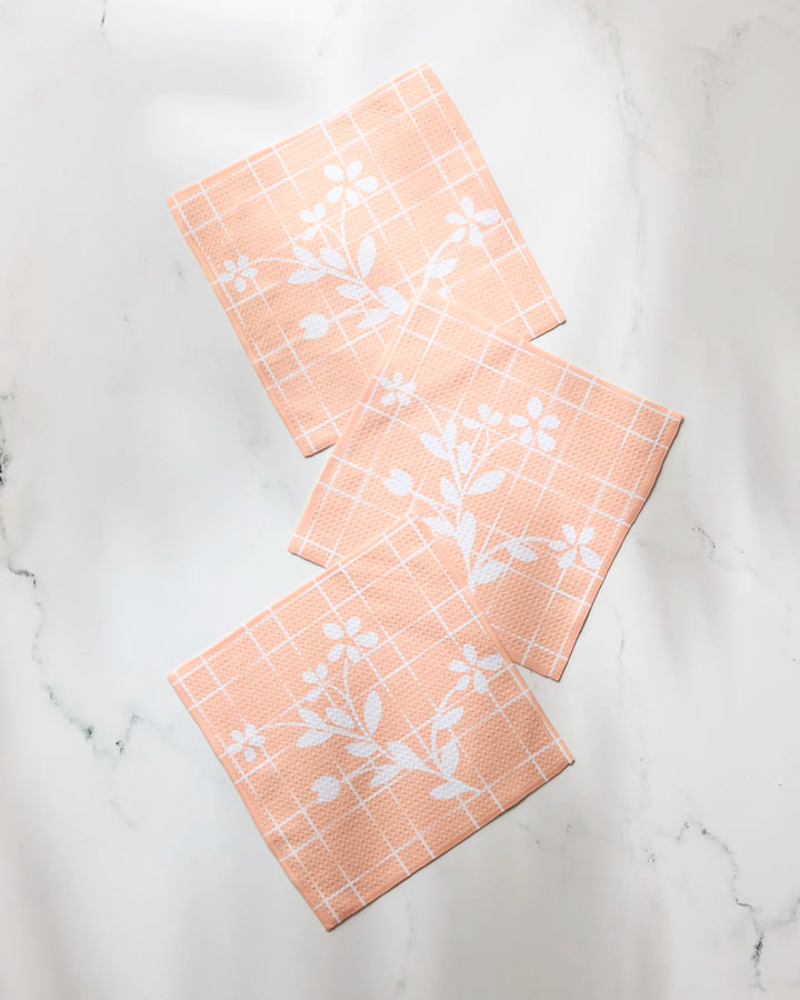 Three peach-colored Quiet Flower cloth napkins by Geometry, featuring a white grid and floral design from their super-absorbent dishtowel set, are arranged on a light marble surface.