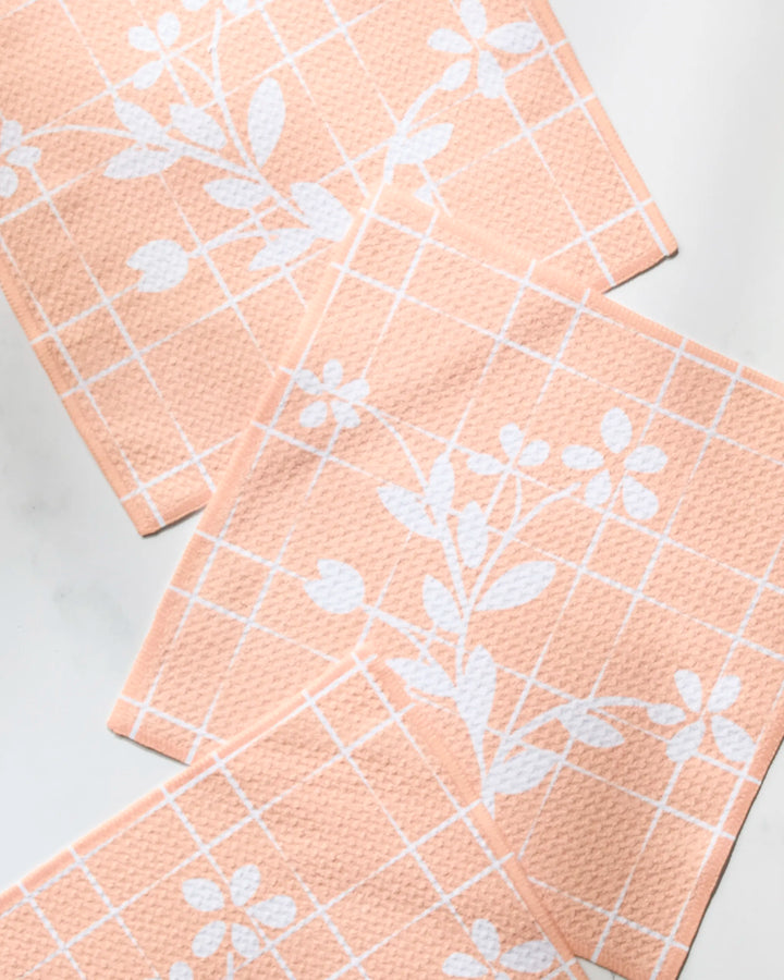 Three Quiet Flower dishcloths by Geometry, featuring a peach color with white grid and floral design made from recycled materials, are arranged in an overlapping layout on a white surface.