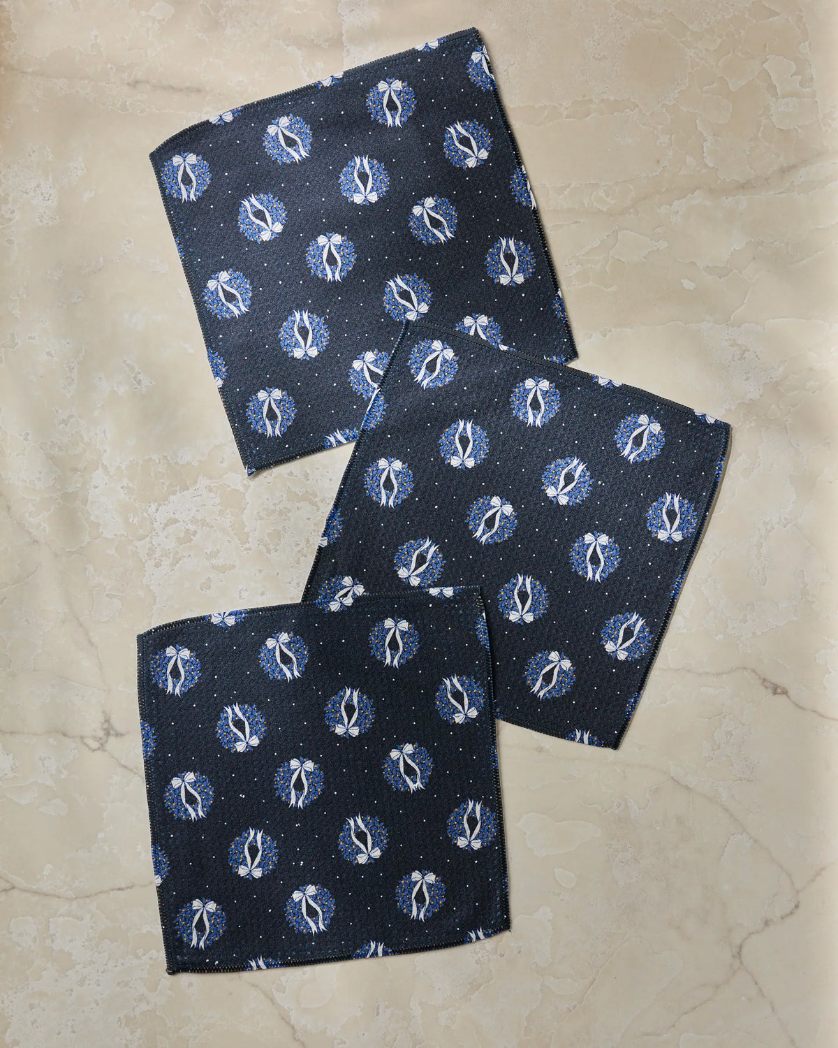 Three “Quiet Skies” hand towels by Geometry, featuring a repeating white and blue circular pattern on navy blue fabric, are displayed on a beige marble surface—offering an elegant dishcloth set that blends stylish design with the practical absorbency of premium kitchen towels.