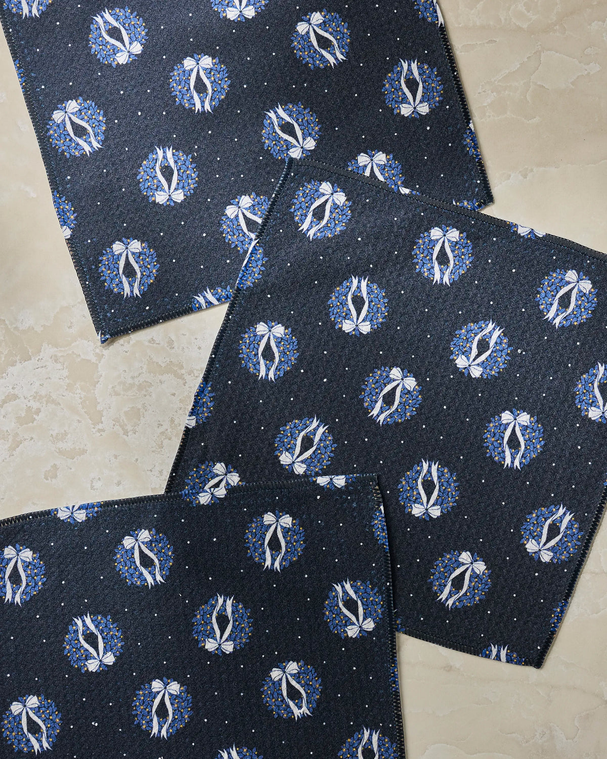 Three “Quiet Skies” cloth napkins by Geometry, showcasing white floral motifs and blue circular accents on a dark blue background, are artfully arranged on a beige marbled surface.