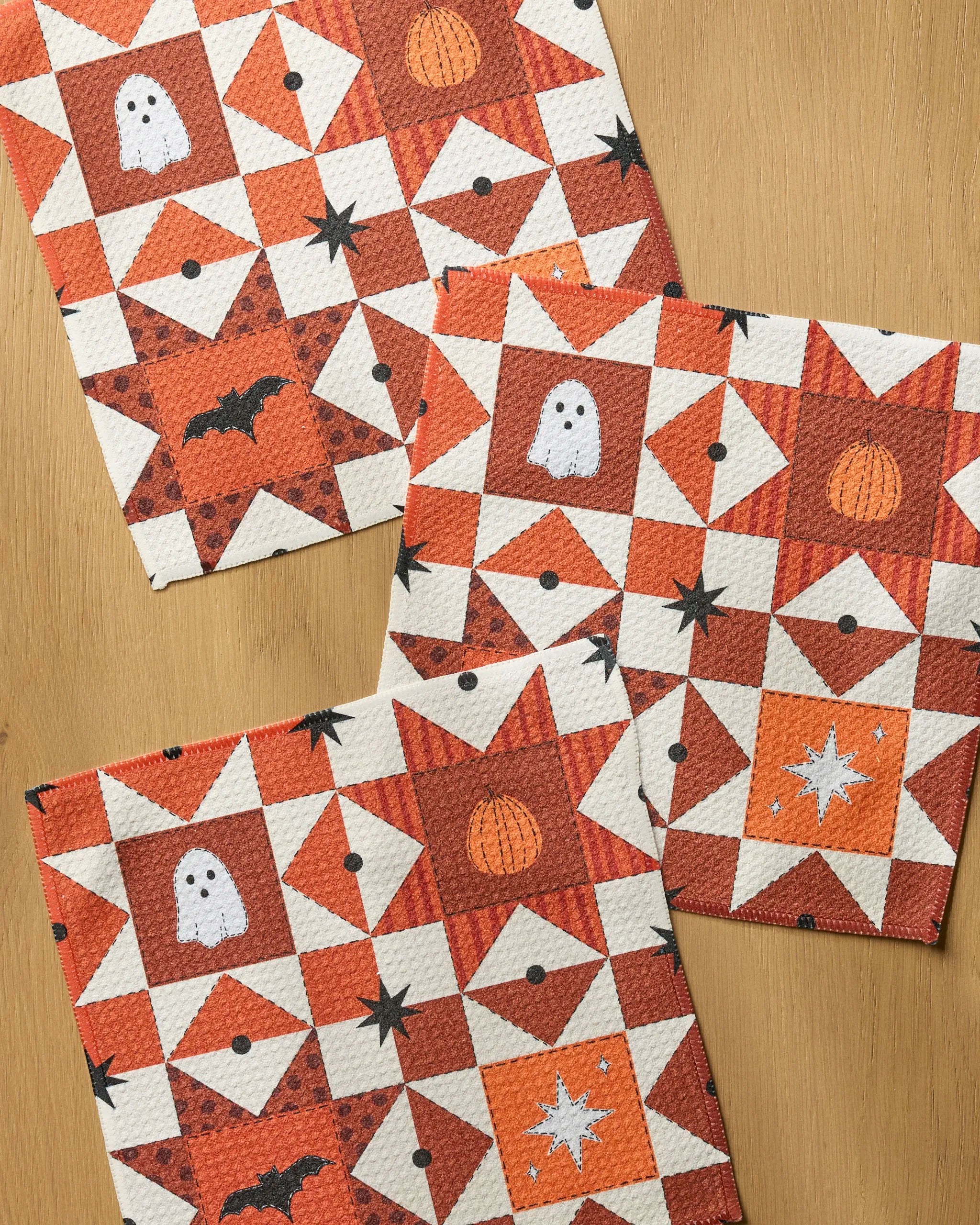 Quilt Work by Geometry: three Halloween-themed quilted squares crafted from recycled materials, featuring orange stars, black bats, white ghosts, pumpkins, and sparkling stars on a wooden surface.