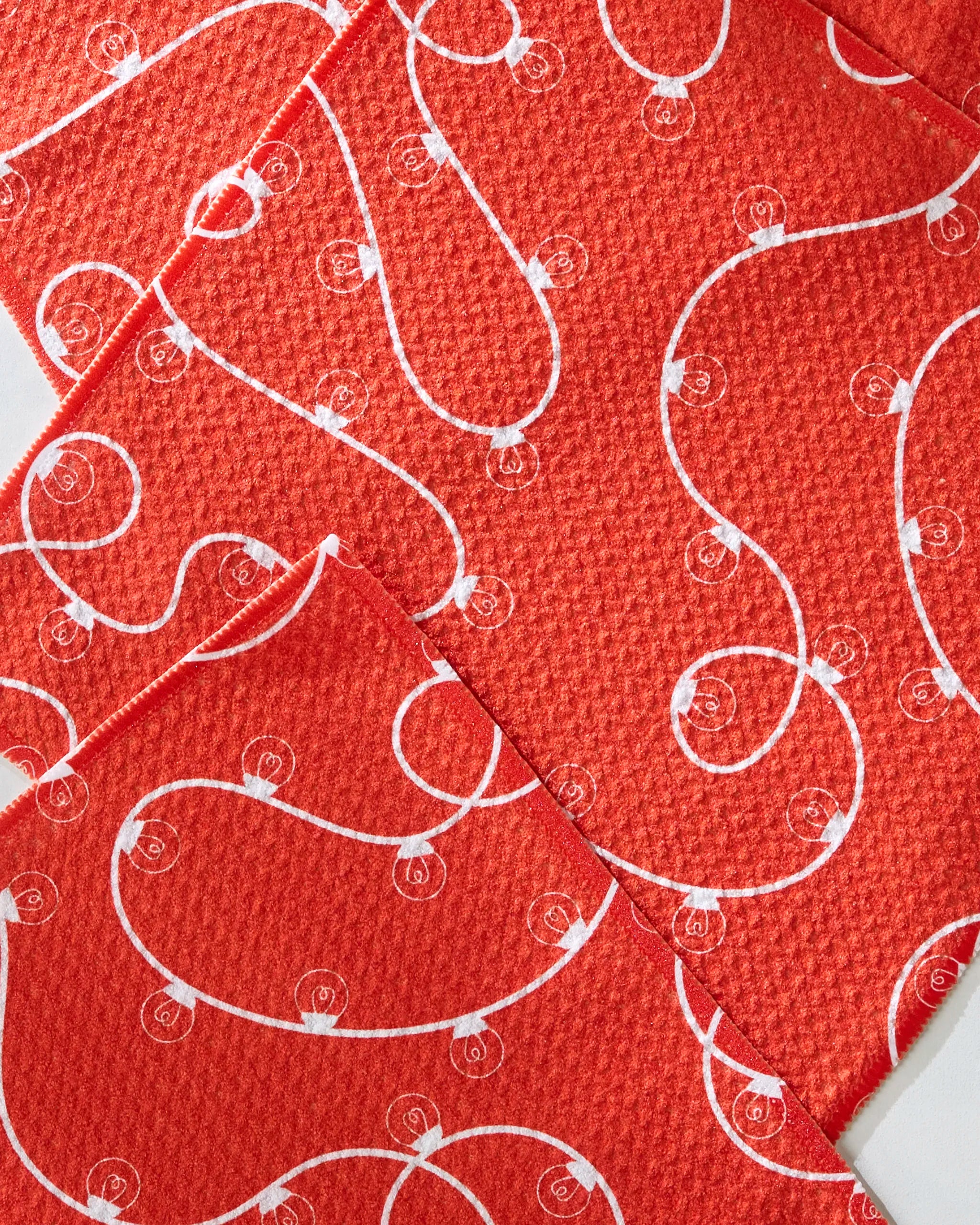 Geometry’s Red Holiday Lights napkins feature overlapping red fabric with textured surfaces, adorned with white swirl patterns and dotted designs, all crafted from recycled materials for an eco-friendly, festive look.