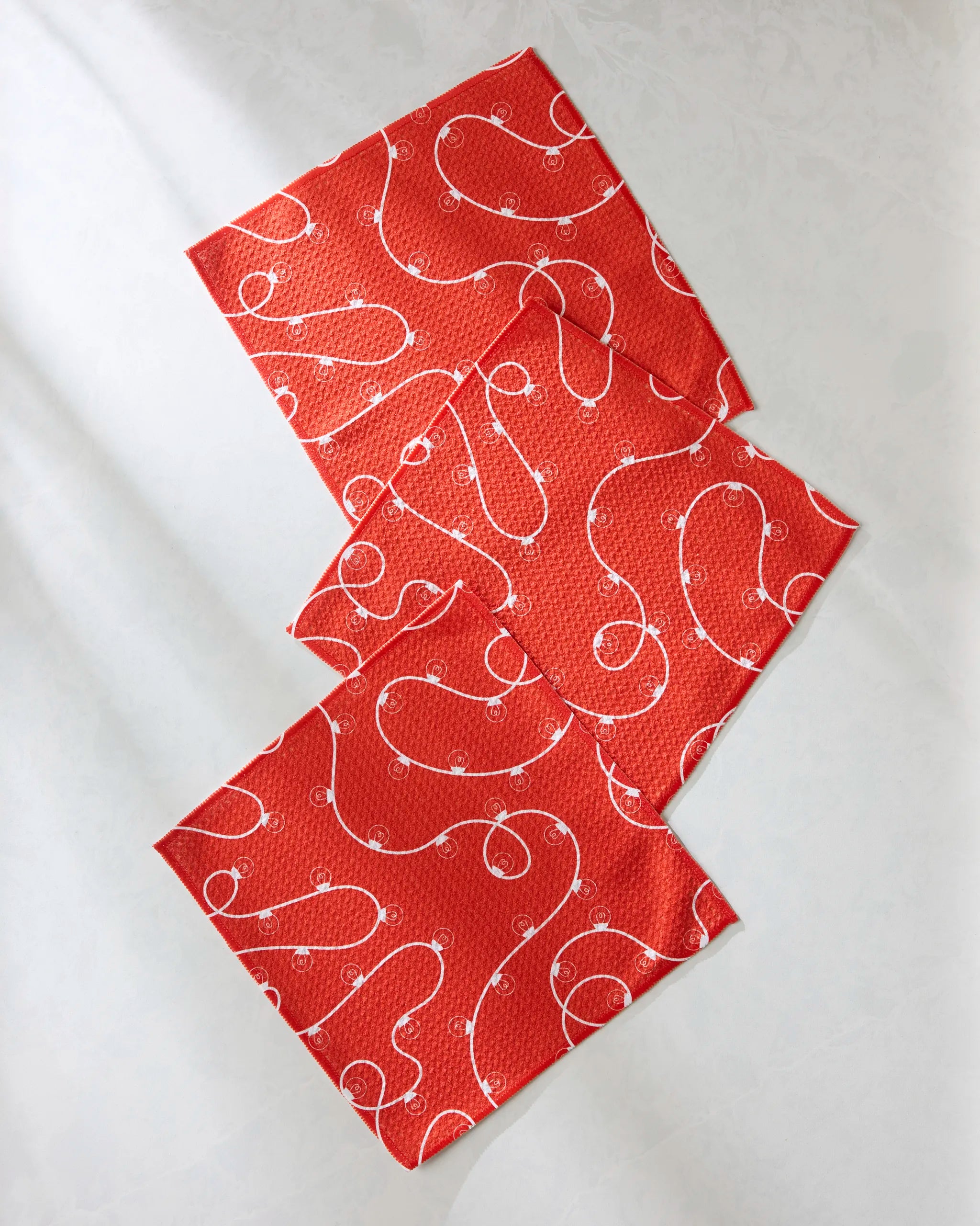 Three "Red Holiday Lights" cloth napkins by Geometry, featuring a festive swirl and dot print, are stylishly layered in a diagonal display on a light surface—perfect as a cheerful kitchen dishcloth set.