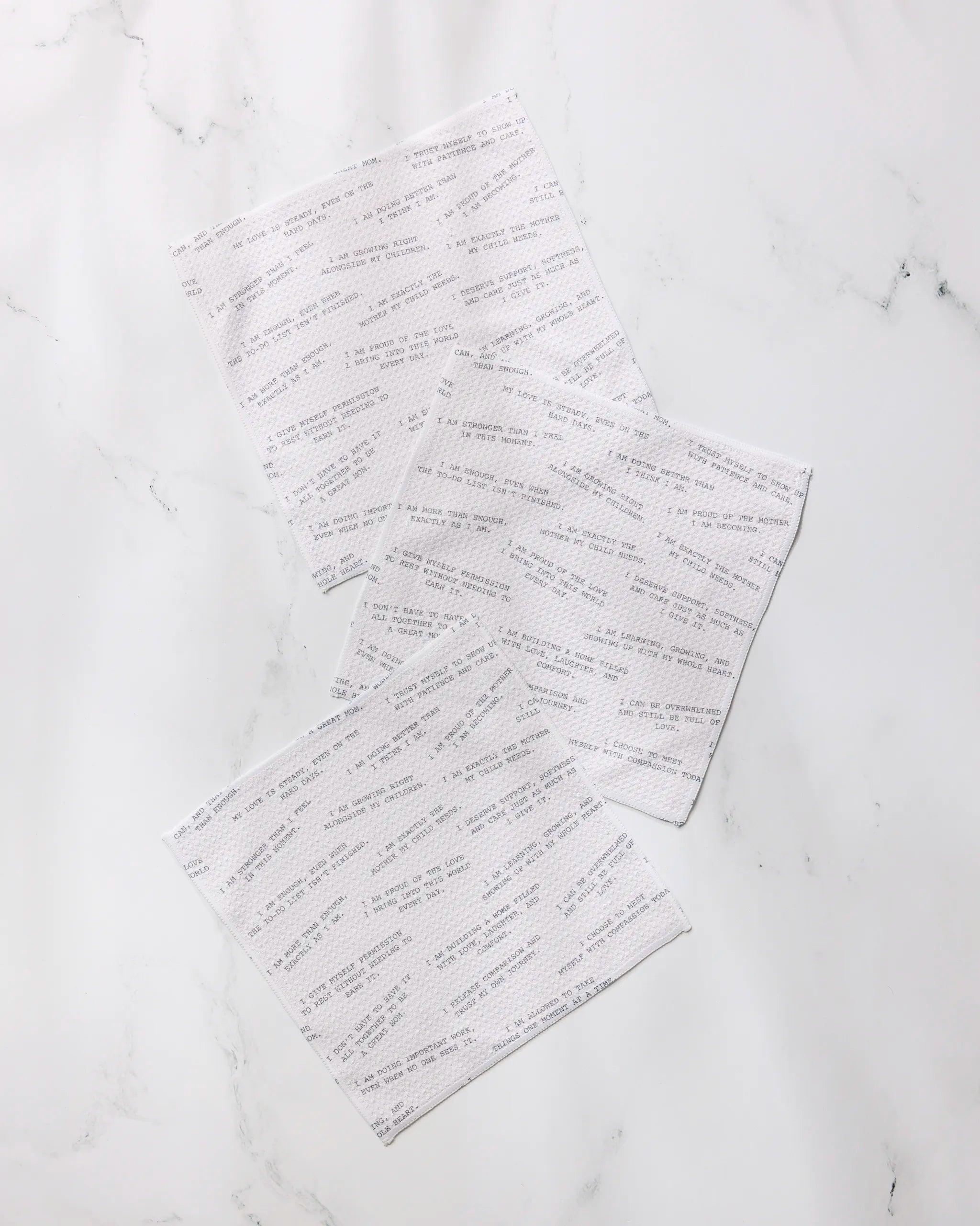 Three "Reminders for Mom" napkins by Geometry, featuring printed gray text on white recycled fabric, are scattered on a white marble surface.