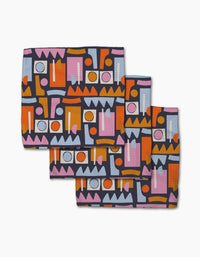 Three folded Retro Shapes Fiesta napkins by Claire Iglesias, made from Geoweave fabric, feature a bold abstract geometric print in orange, pink, white, light blue, and purple on dark blue and are arranged overlapping on a white surface.