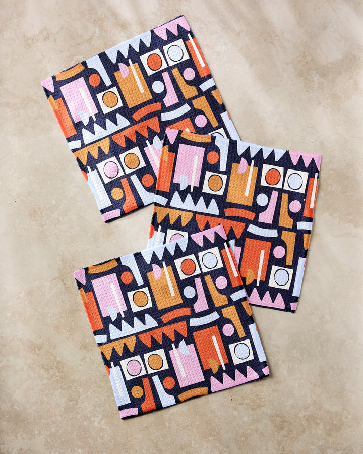 The Claire Iglesias "Retro Shapes Fiesta" set features three rectangular dishcloths with bold geometric patterns in pink, orange, white, and black—super-absorbent and crafted from recycled materials.
