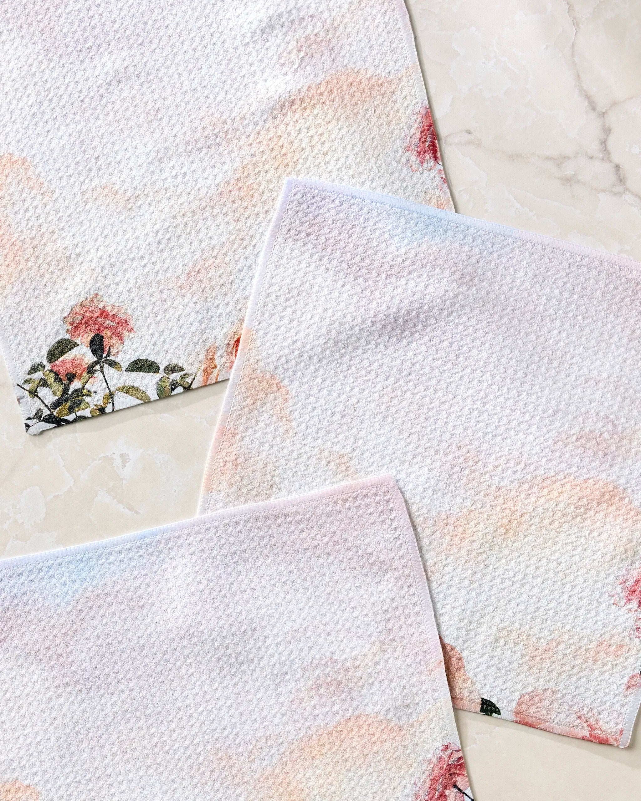 The Stefanie Bales Rhapsody in Bloom set includes three recycled dishcloths in soft pastels with floral designs, displayed on marble. The upper left cloth showcases the most prominent flowers, blending eco-friendly function with style.