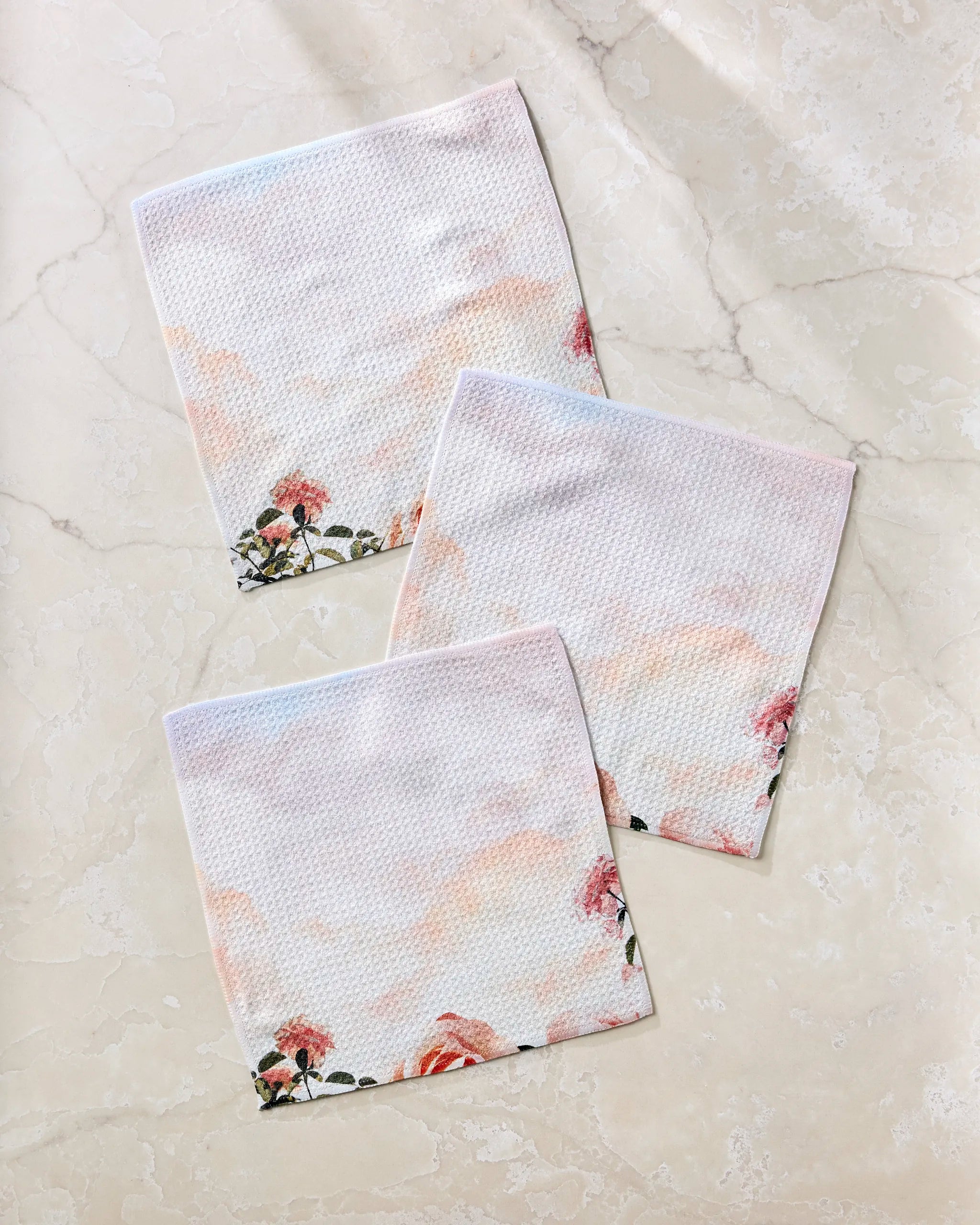 Three pastel recycled dishcloths from Stefanie Bales’ Rhapsody in Bloom collection, featuring pink and orange floral designs and a subtle textured pattern, are arranged overlapping on a light beige marble surface for a relaxed look.