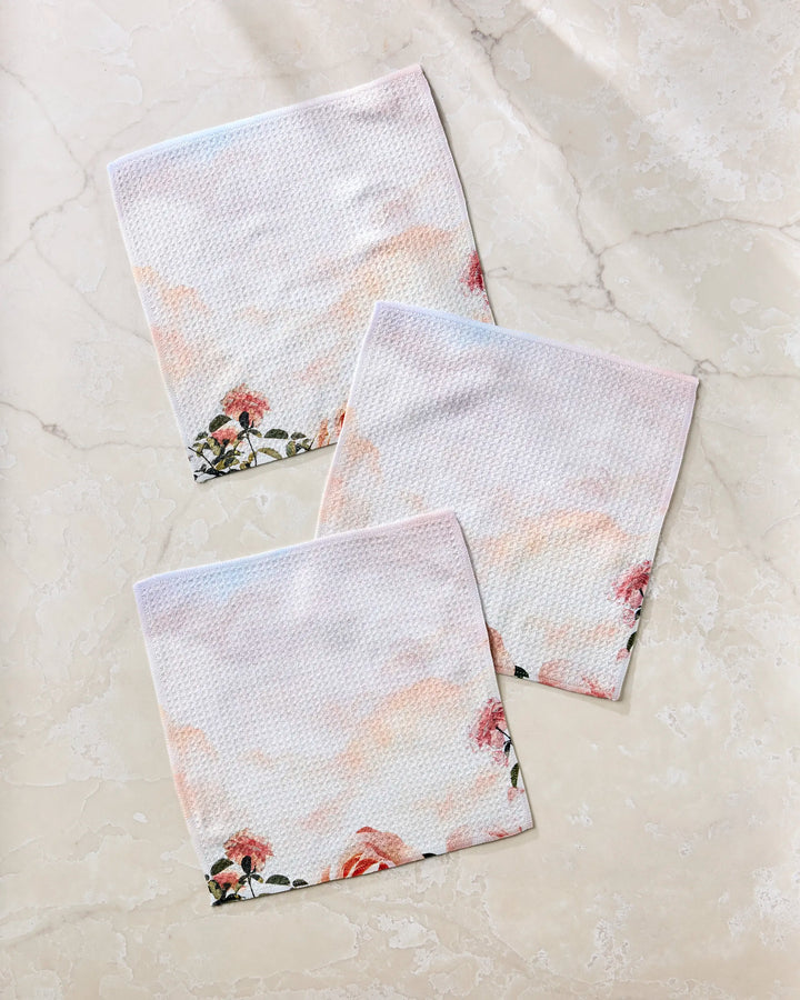 Three pastel recycled dishcloths from Stefanie Bales’ Rhapsody in Bloom collection, featuring pink and orange floral designs and a subtle textured pattern, are arranged overlapping on a light beige marble surface for a relaxed look.