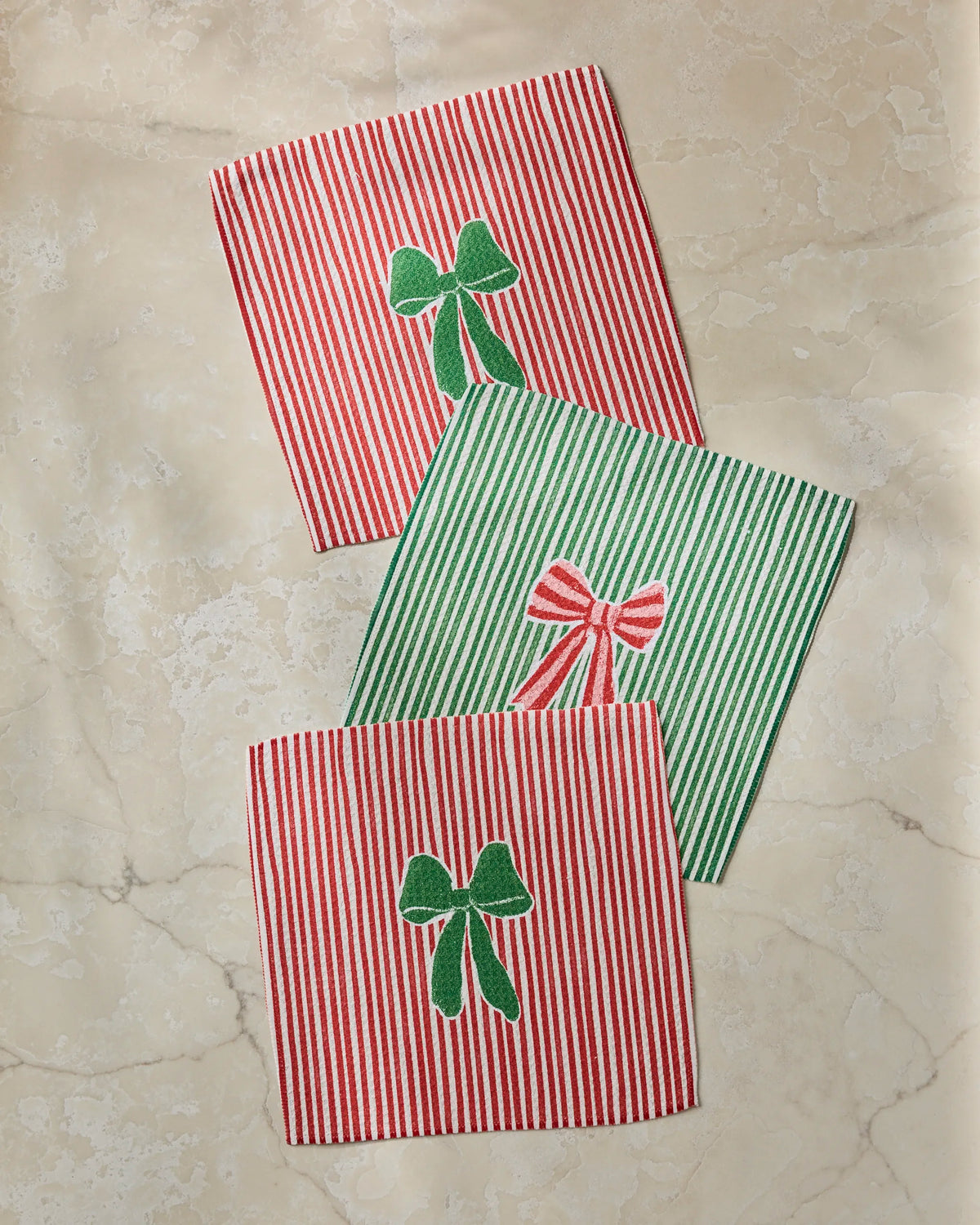 Three “Ribbon Bows” cloth napkins by Geometry—two featuring red stripes with embroidered green bows, and one with green stripes and a red bow—are artfully arranged on a light marble surface, creating a delightful set that’s perfect for any occasion. These absorbent kitchen towels combine festive charm with daily practicality.