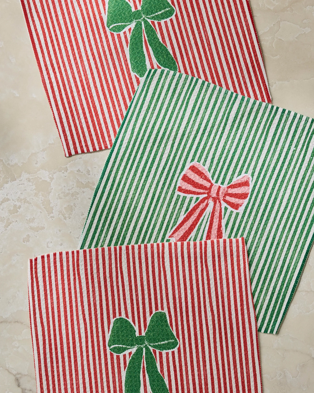 Three Ribbon Bows napkins from Geometry—two featuring red and white stripes with green ribbon bows, and one in green and white stripes with a red bow—are arranged on a marble surface, creating a festive set perfect for any gathering.