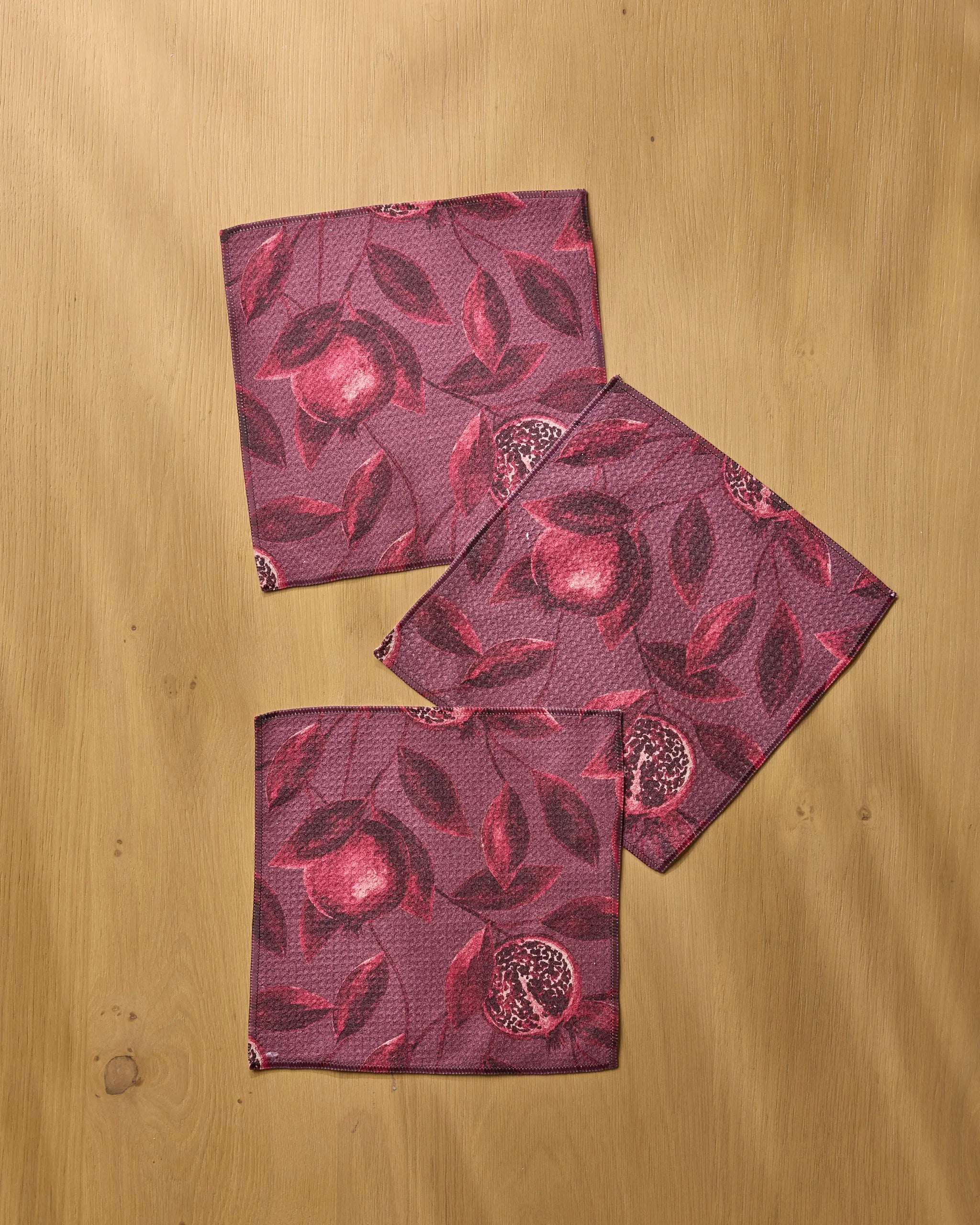 Three square, purple Rich Garnet napkins by Geometry, made from recycled materials, feature pomegranate and leaf designs. Arranged on a light wood surface, they overlap to showcase the eco-friendly set's vibrant fruit patterns.